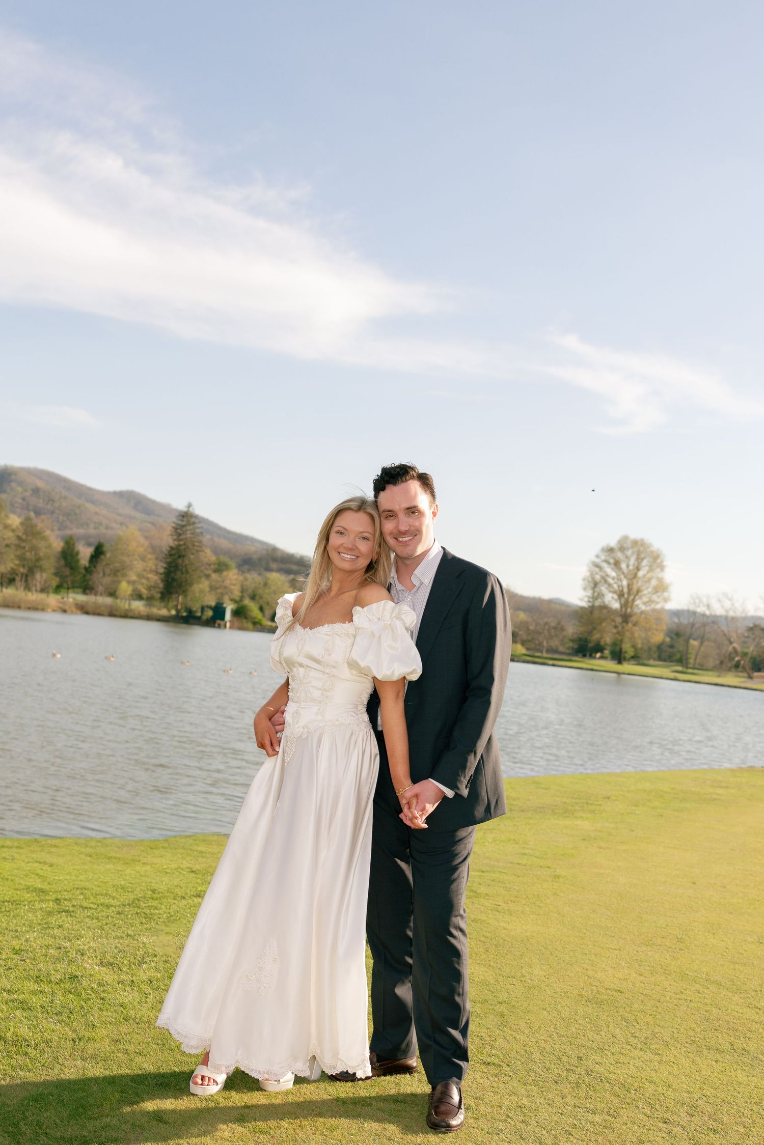 Bride in white off-shoulder gown and groom in dark suit hold hands by a lake with mountain views.