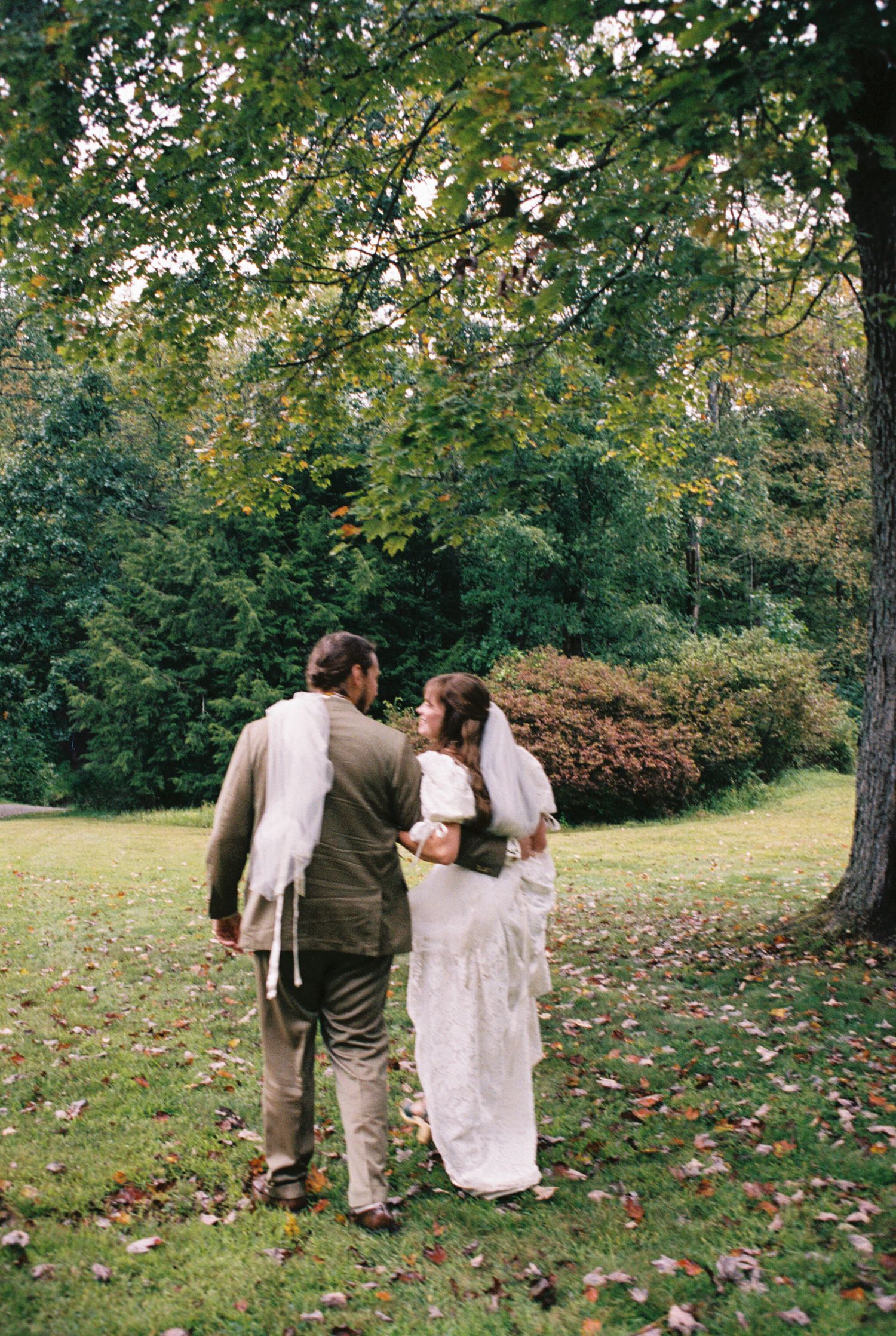 A couple in wedding attire share a tender moment on a grassy lawn surrounded by autumn trees and fallen leaves.