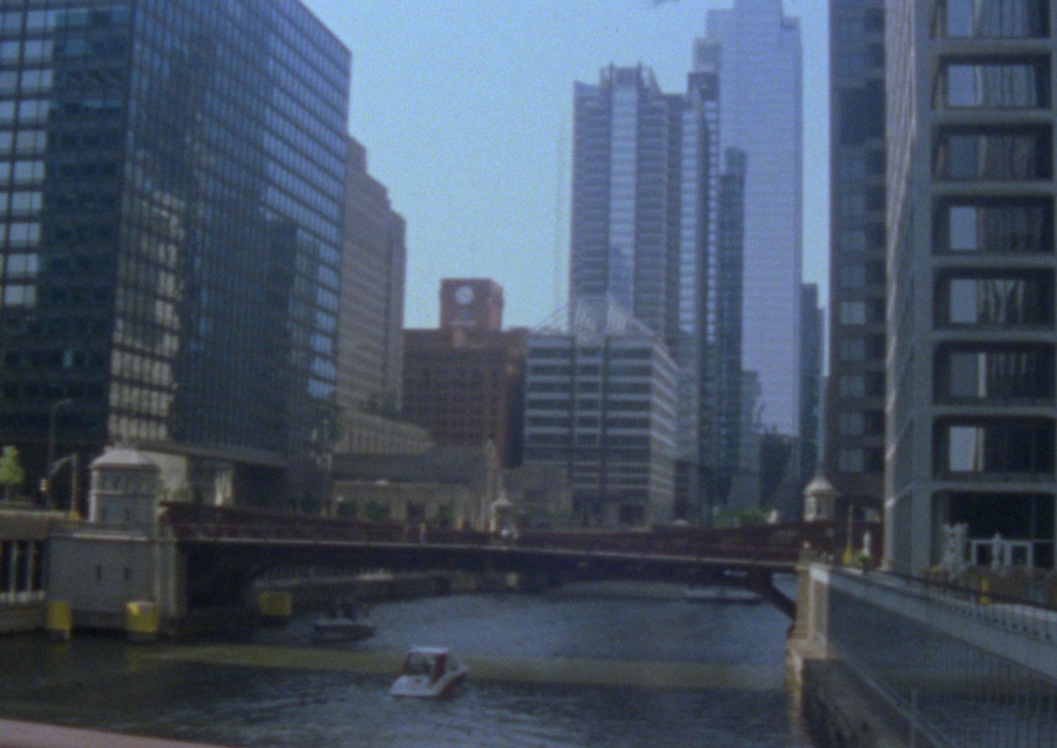 View of Chicago River with tall skyscrapers and a red bridge crossing between buildings on an overcast day.