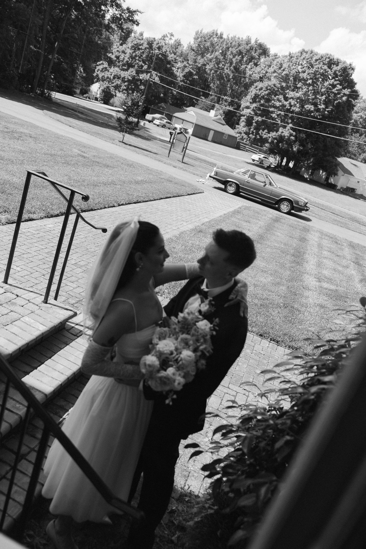 Black and white photo of a bride and groom embracing outdoors, holding a bouquet near brick steps with a vintage car and trees behind.