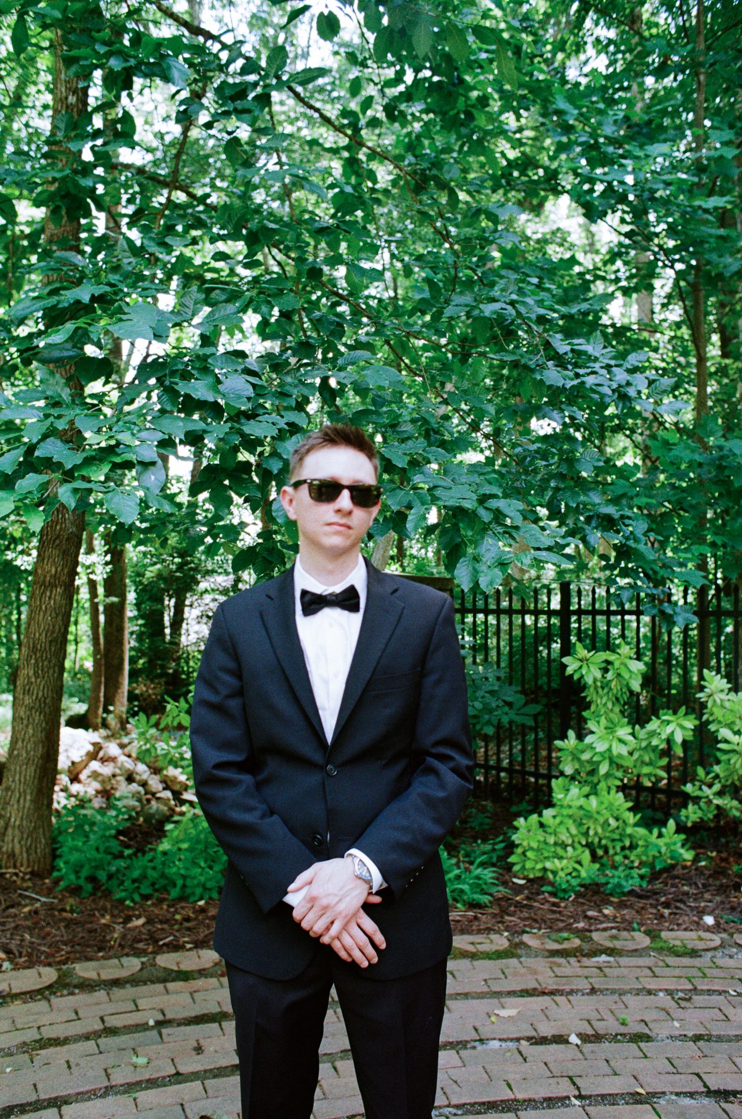 Young man in a black tuxedo, bow tie and sunglasses stands with hands clasped on a brick path beside green trees and a black iron fence.