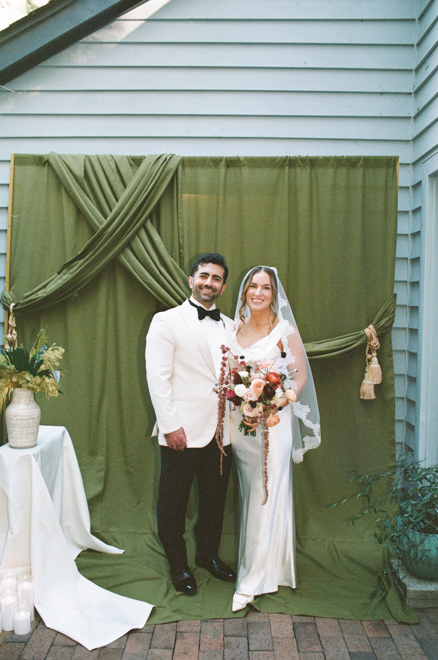 Smiling bride in a veil and groom in a white tuxedo jacket pose with a bouquet before an olive green draped wedding backdrop.