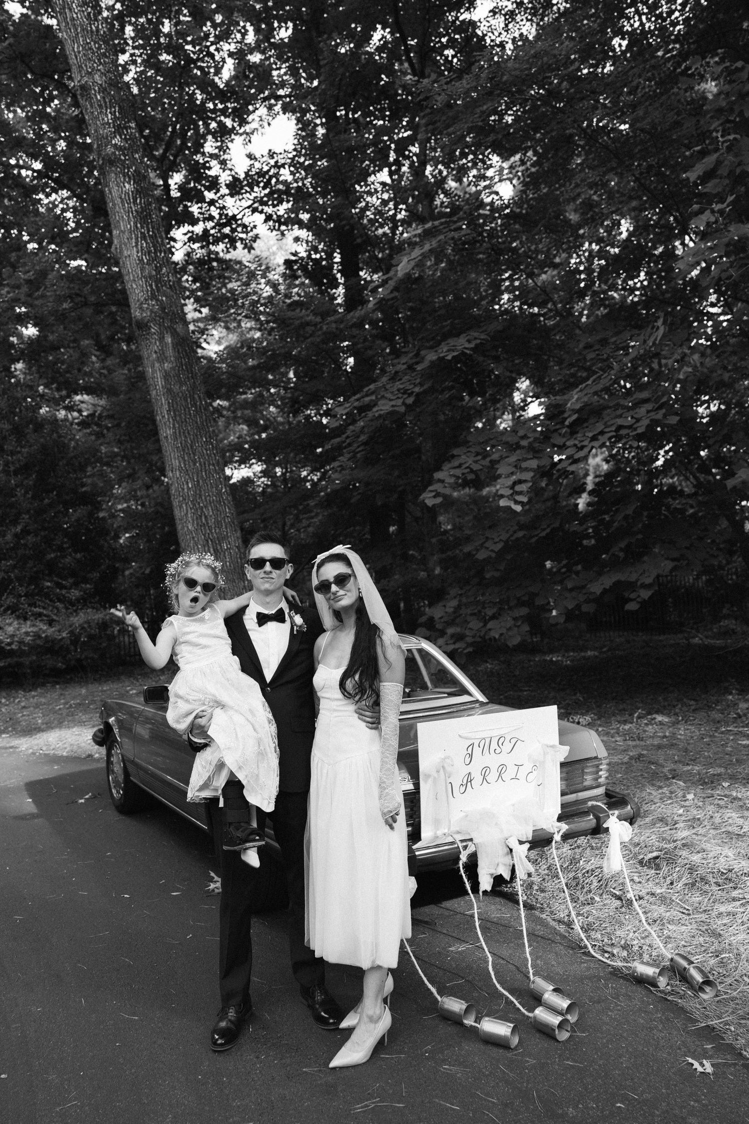 Black and white photo of a just married couple in sunglasses holding a flower girl beside a vintage car with tin cans.