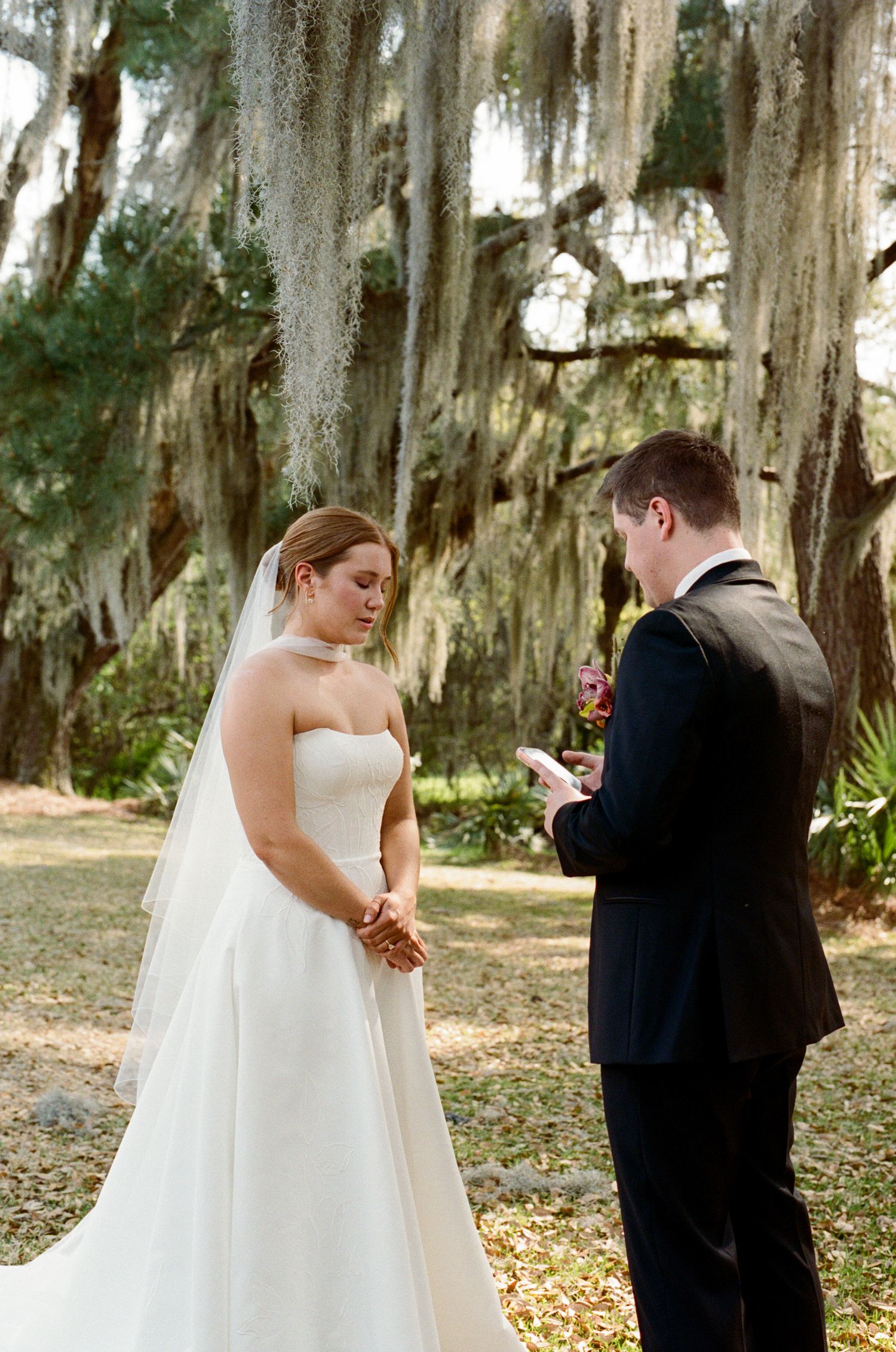 Bride and groom exchange vows outdoors under Spanish moss-draped oak trees during an elegant garden wedding ceremony.