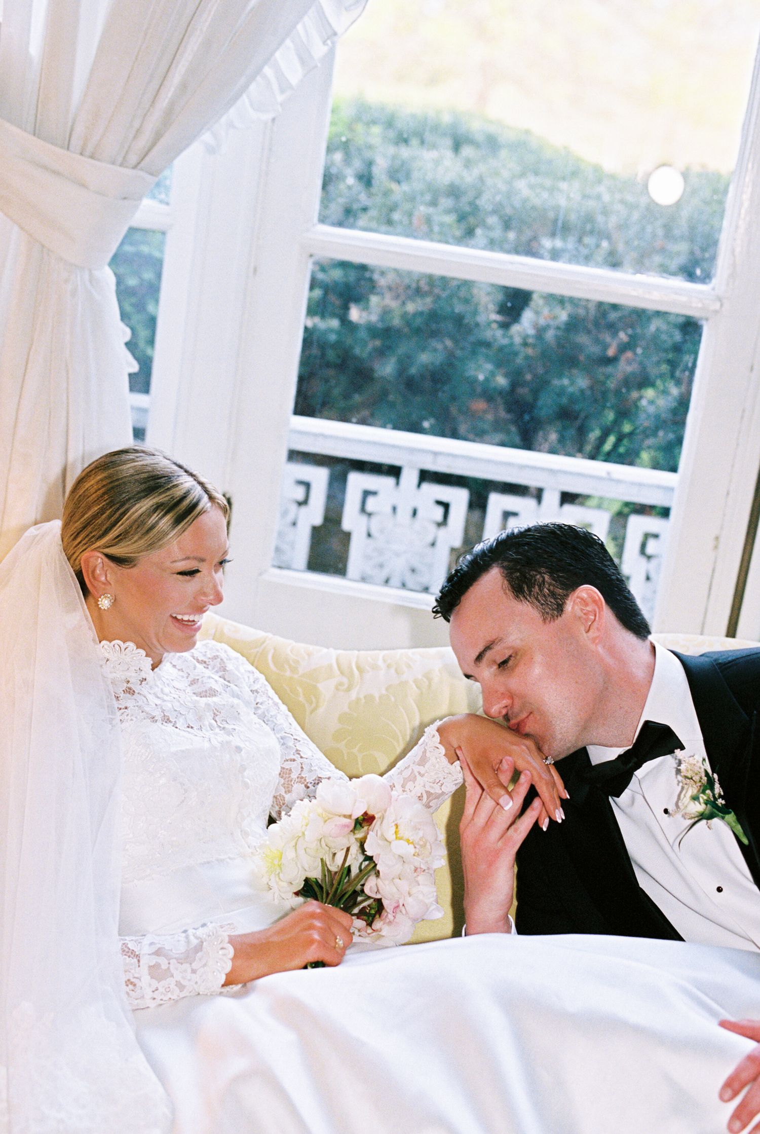Groom in tuxedo kissing bride's hand as she smiles in lace wedding dress, holding white bouquet indoors.