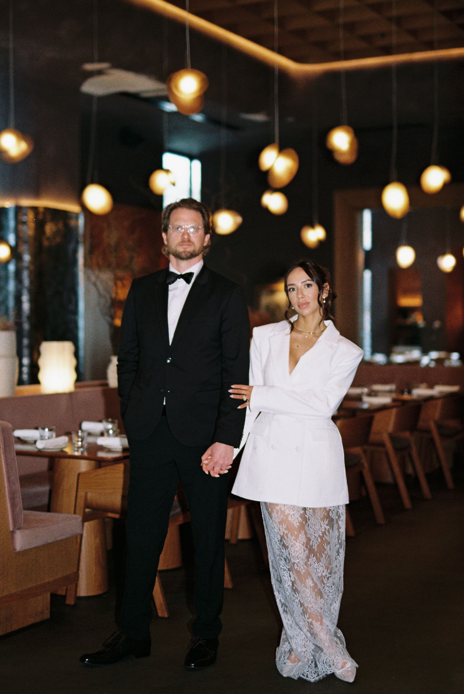 Elegant couple in black tuxedo and white blazer lace skirt posing in upscale restaurant with warm pendant lights.