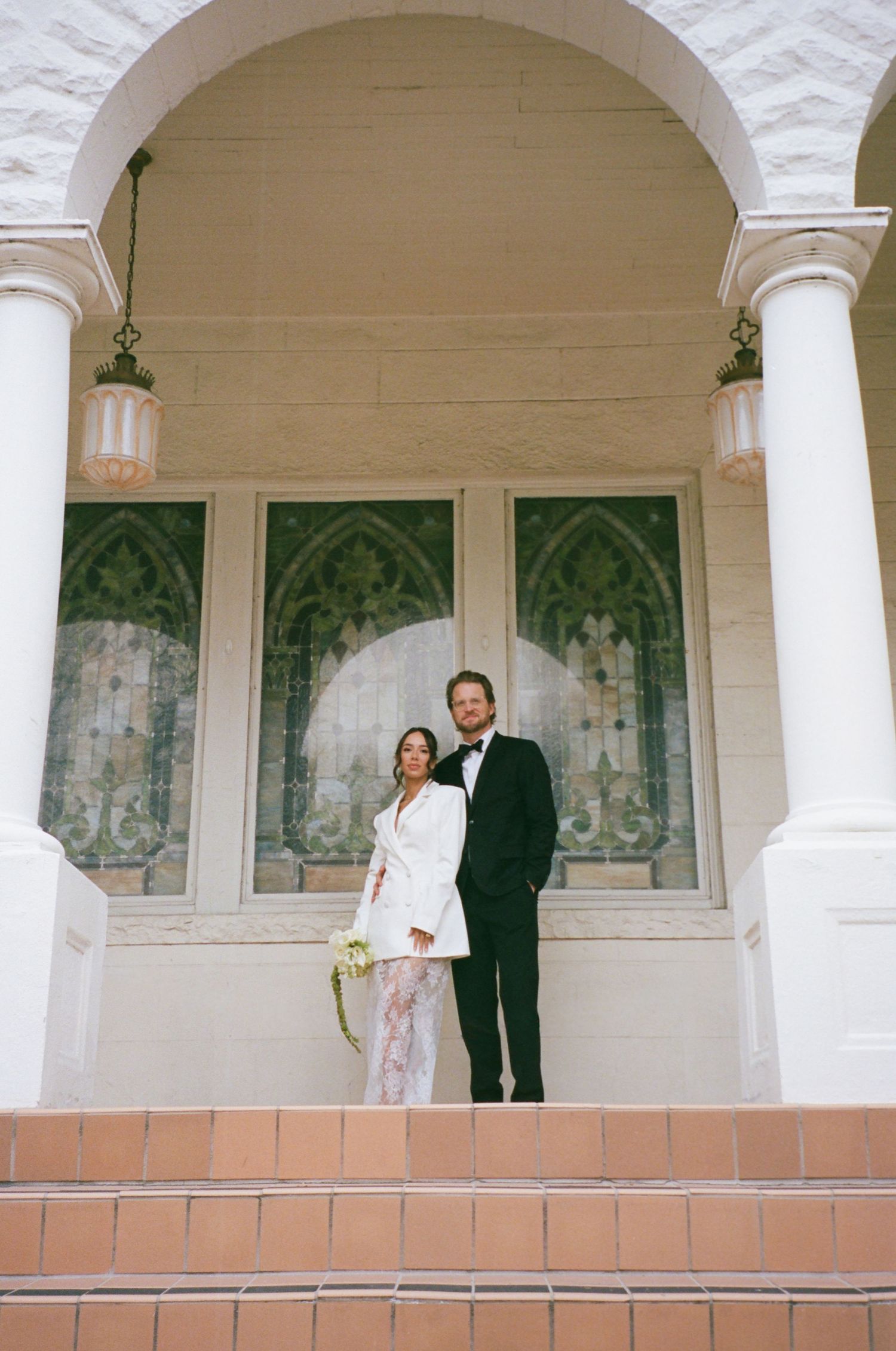 Bride in white blazer and groom in black tuxedo standing on steps of elegant venue with arched columns and stained glass windows.