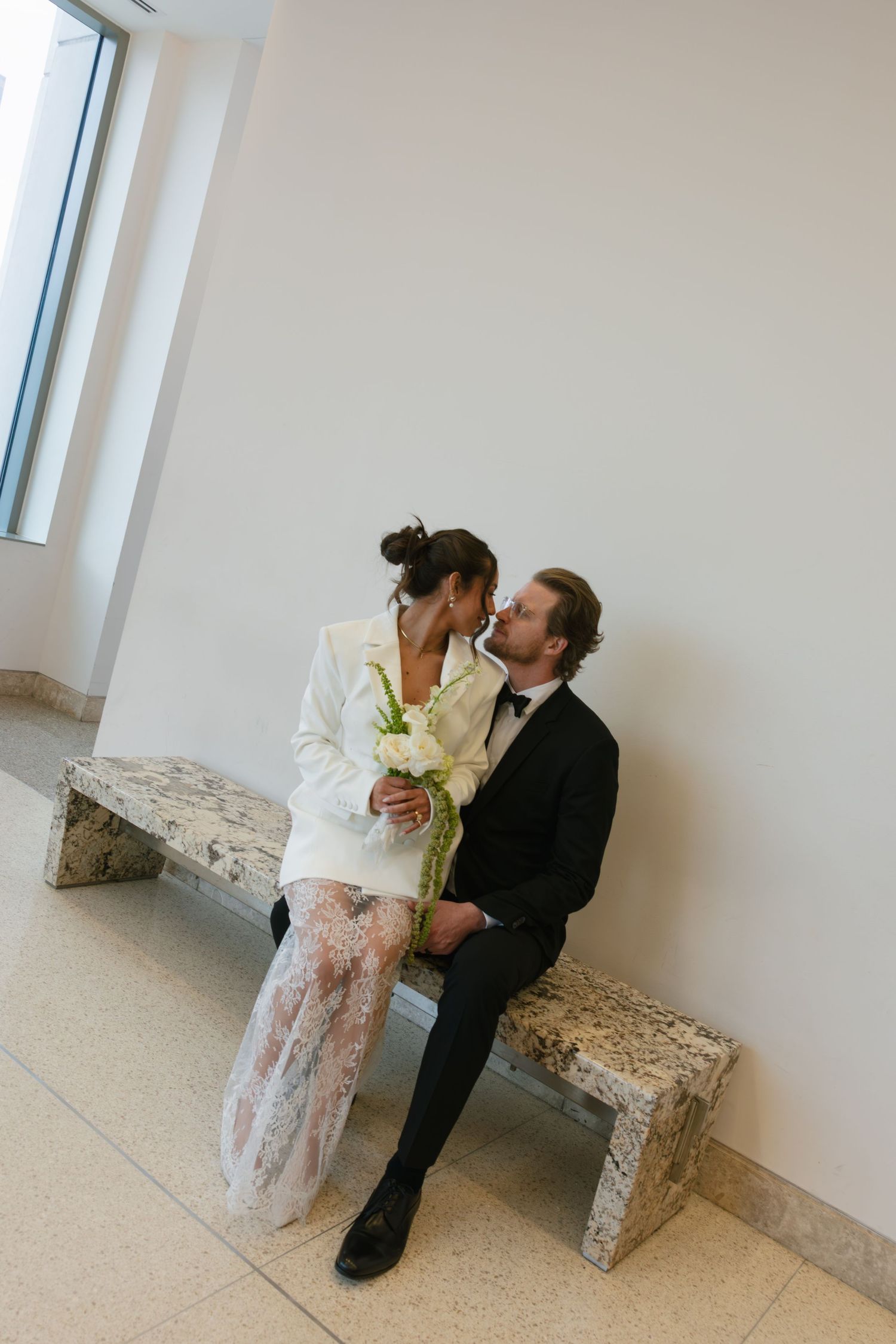 Bride and groom sharing a romantic kiss on a marble bench, bride in white blazer dress holding lily bouquet, groom in black tuxedo.