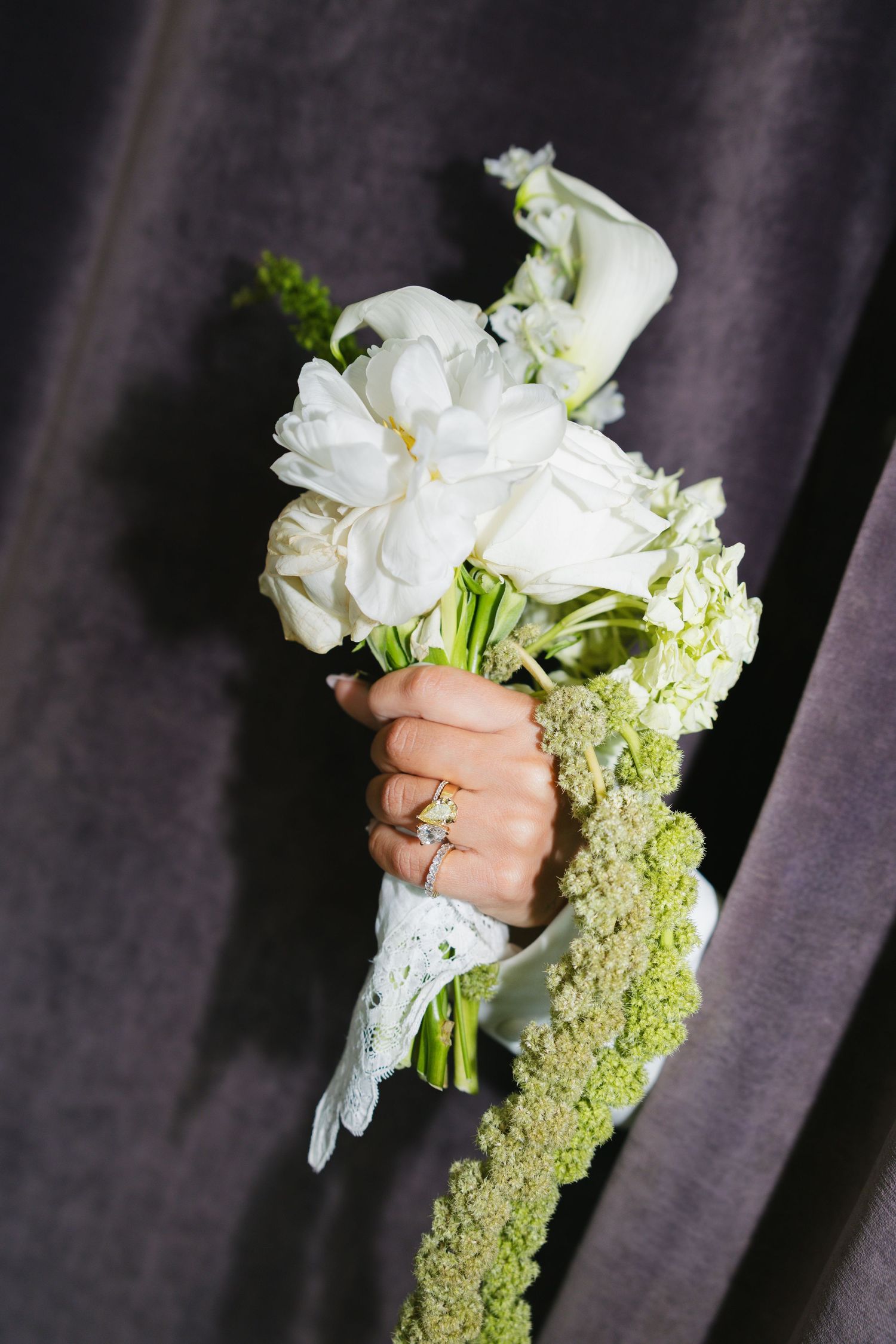 Hand holding a white bridal bouquet with peonies, calla lilies, and green accents, wrapped in lace, against gray fabric.