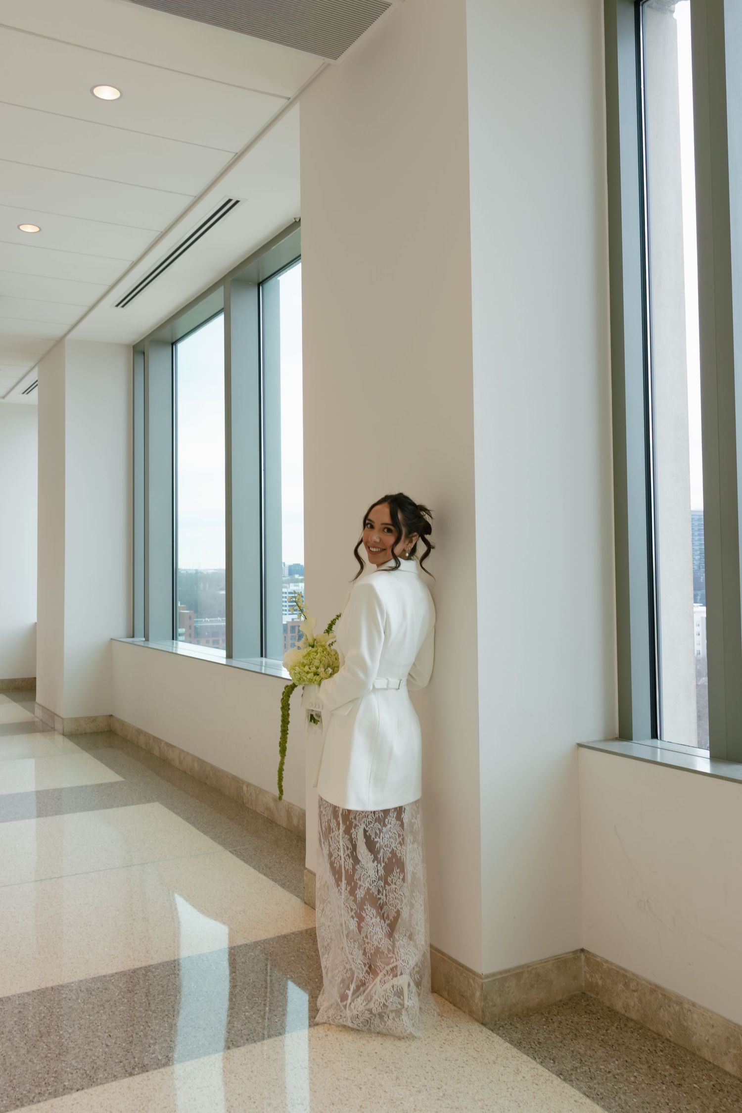 Bride in white blazer and lace skirt holding green bouquet poses in sunlit hallway with large windows.
