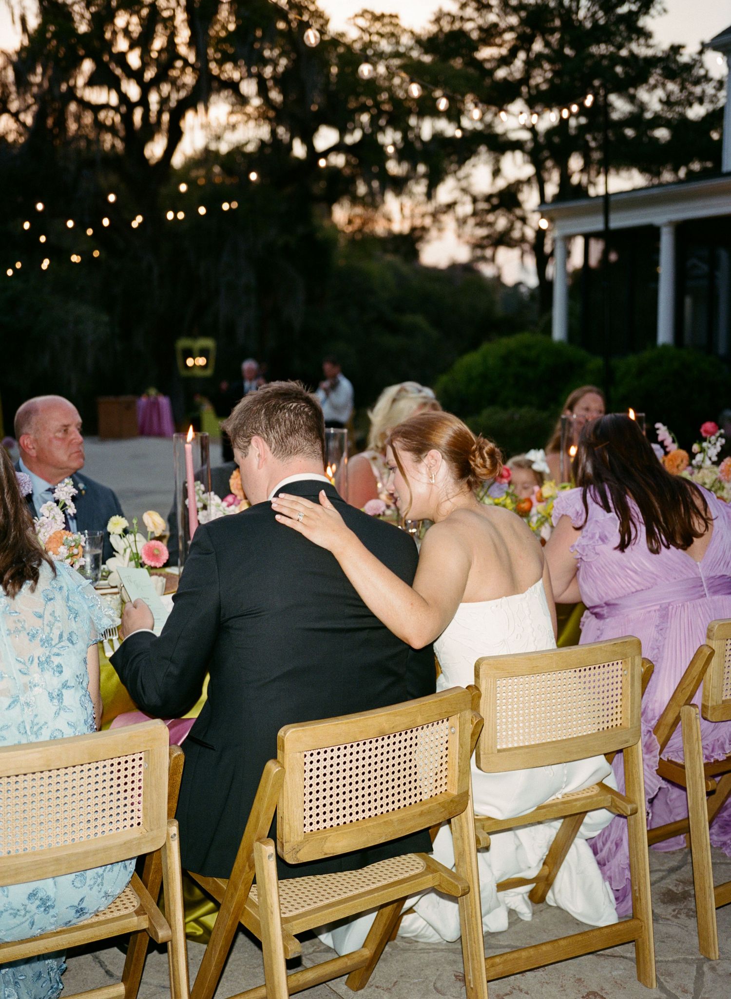 Bride and groom share an intimate moment at outdoor wedding reception with string lights and colorful floral centerpieces.