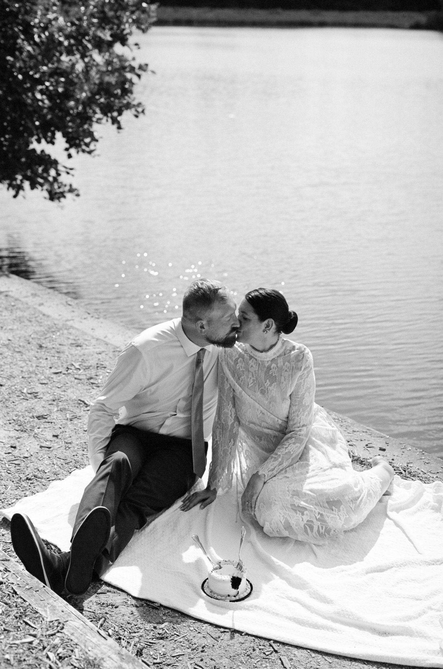 Black and white photo of a couple sharing a kiss while sitting on a blanket by a tranquil lake on their wedding day.