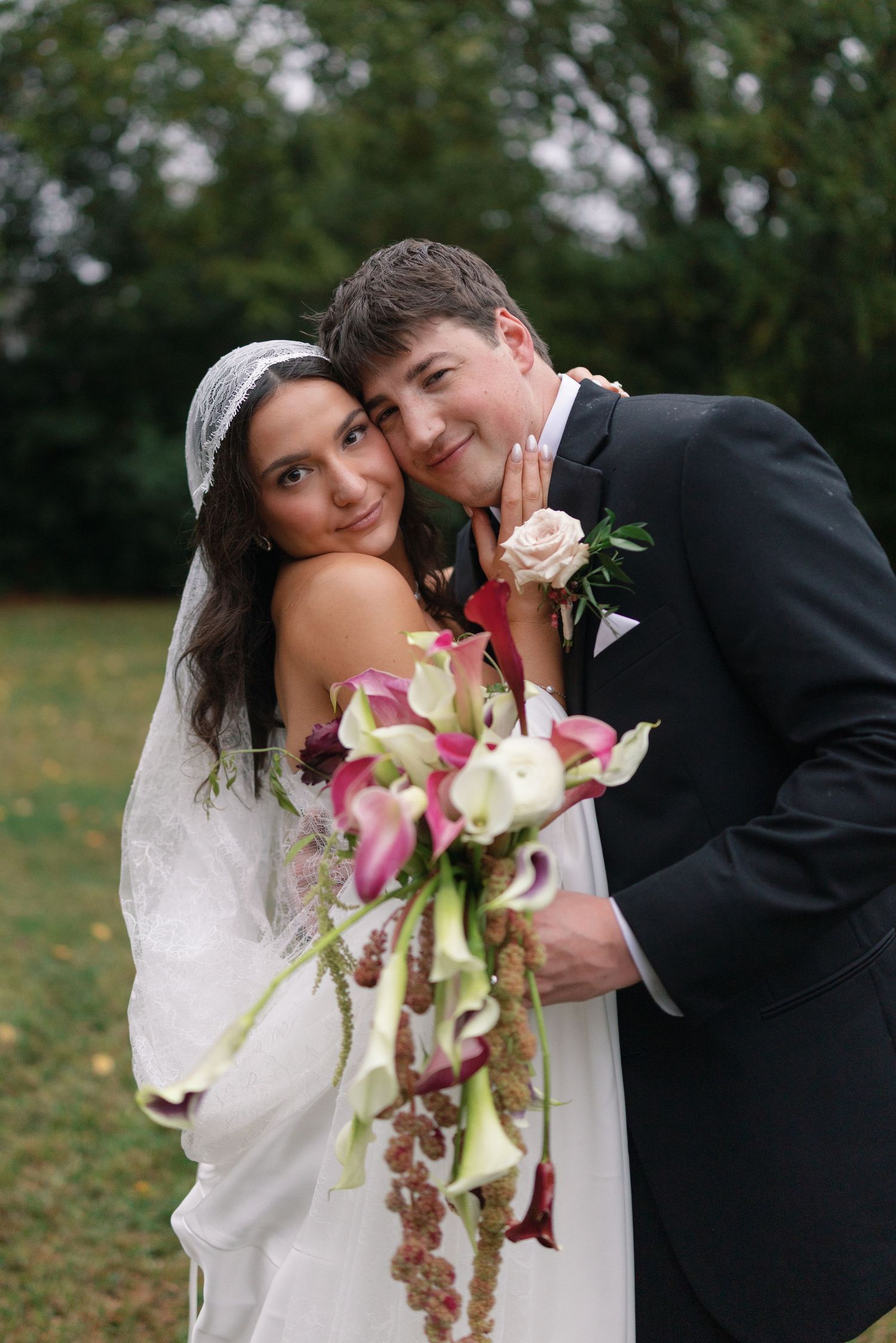 Smiling bride in a lace veil and groom in a dark suit embrace outdoors, holding a cascading bouquet of calla lilies and burgundy blooms.