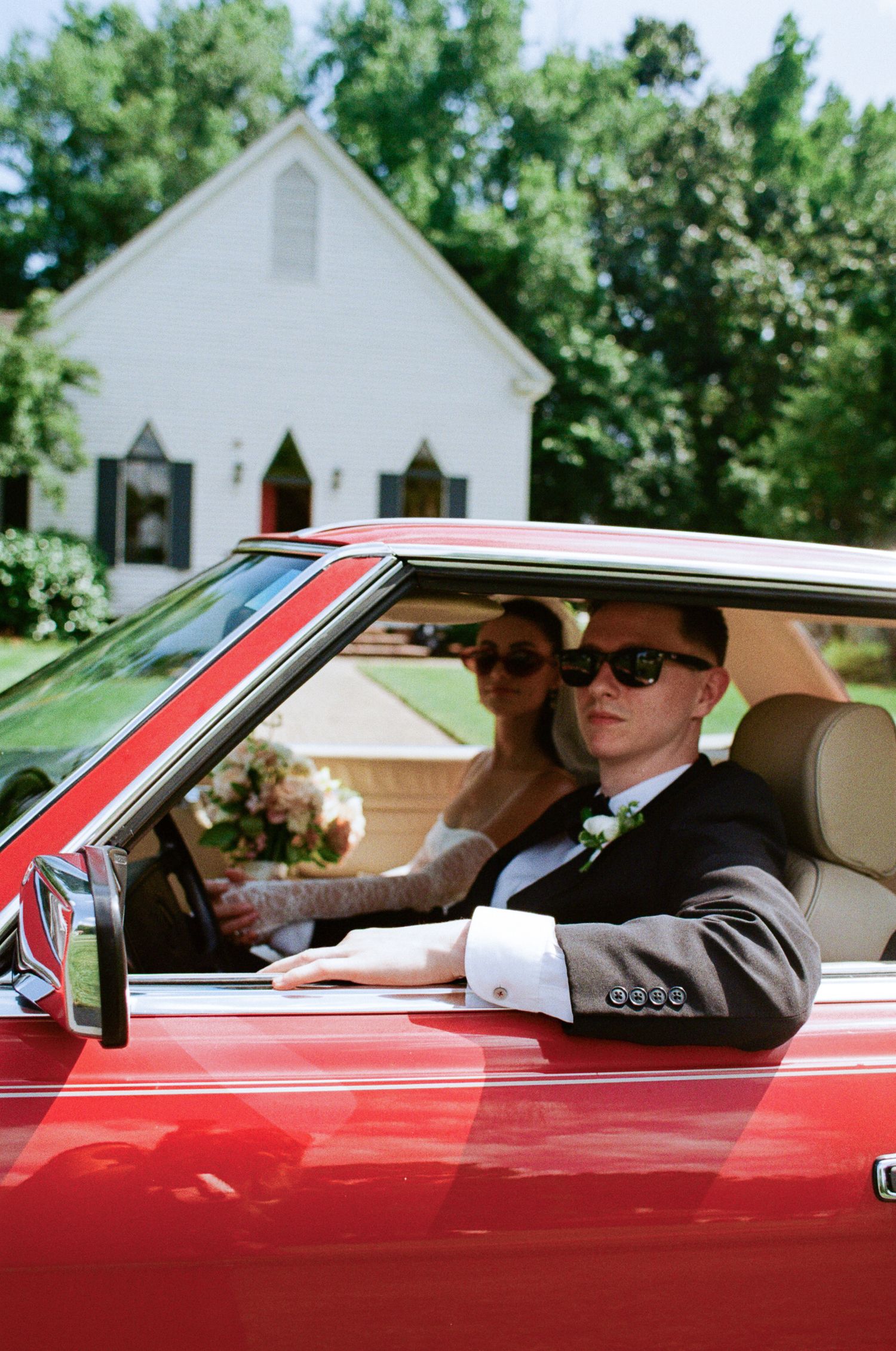 Groom in a dark suit and sunglasses sits in a red vintage car beside a bride holding a bouquet, with a white chapel behind them.