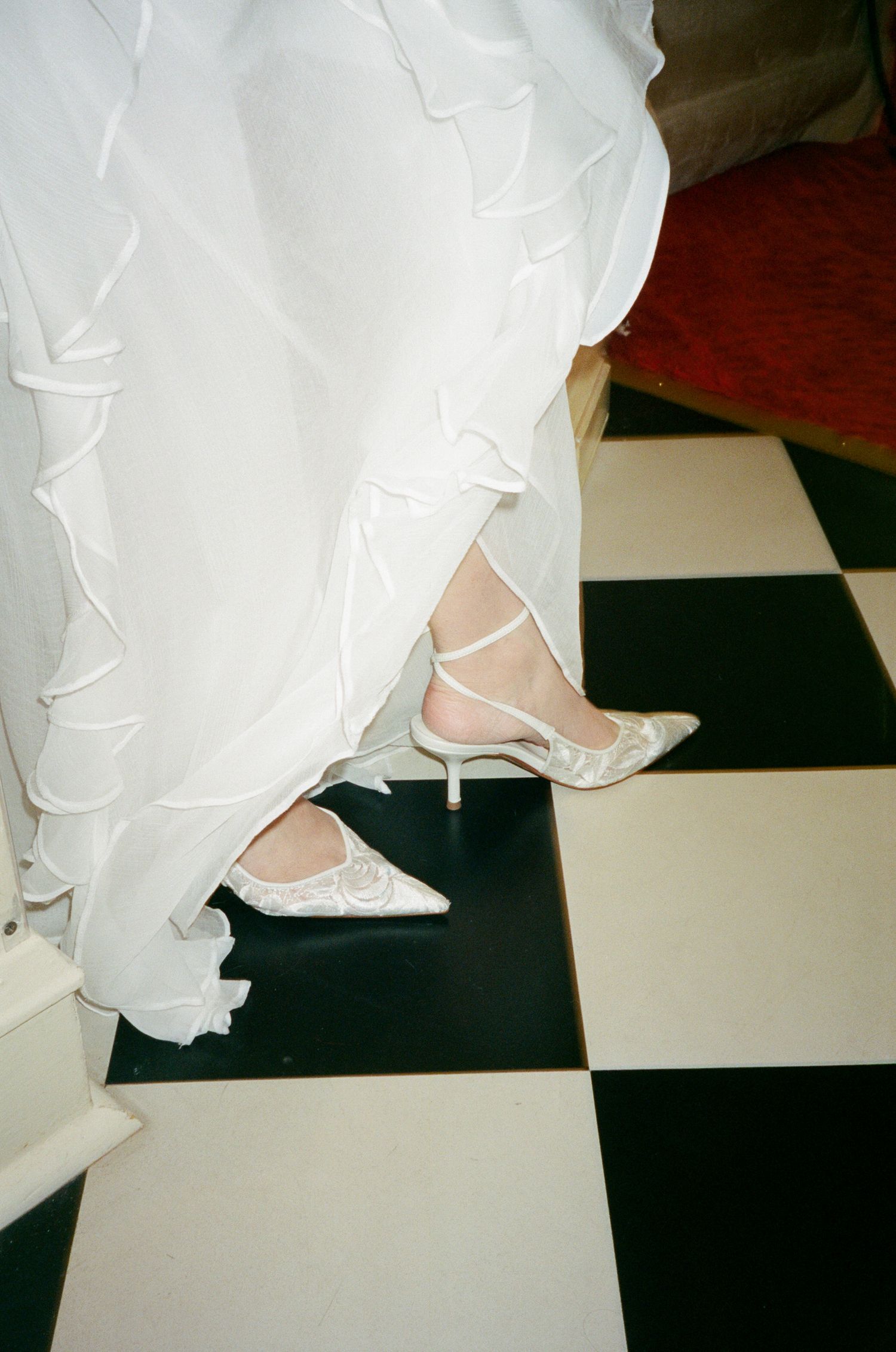 White bridal shoes with straps and heels shown on a black and white checkered floor beneath flowing wedding dress fabric.