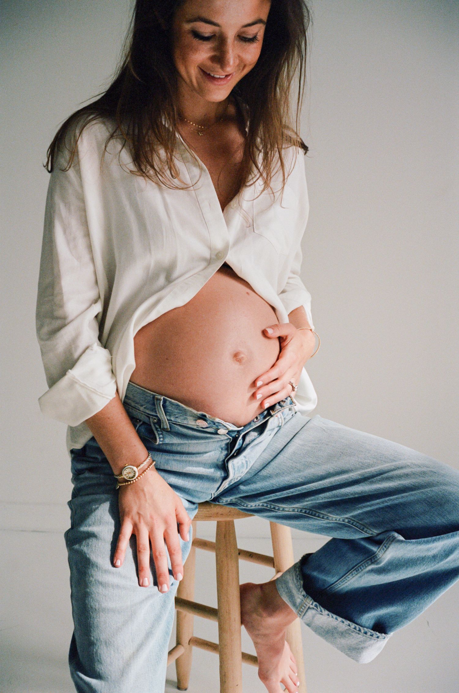 Person wearing a white shirt and loose jeans sits casually on a wooden stool in a minimalist setting.