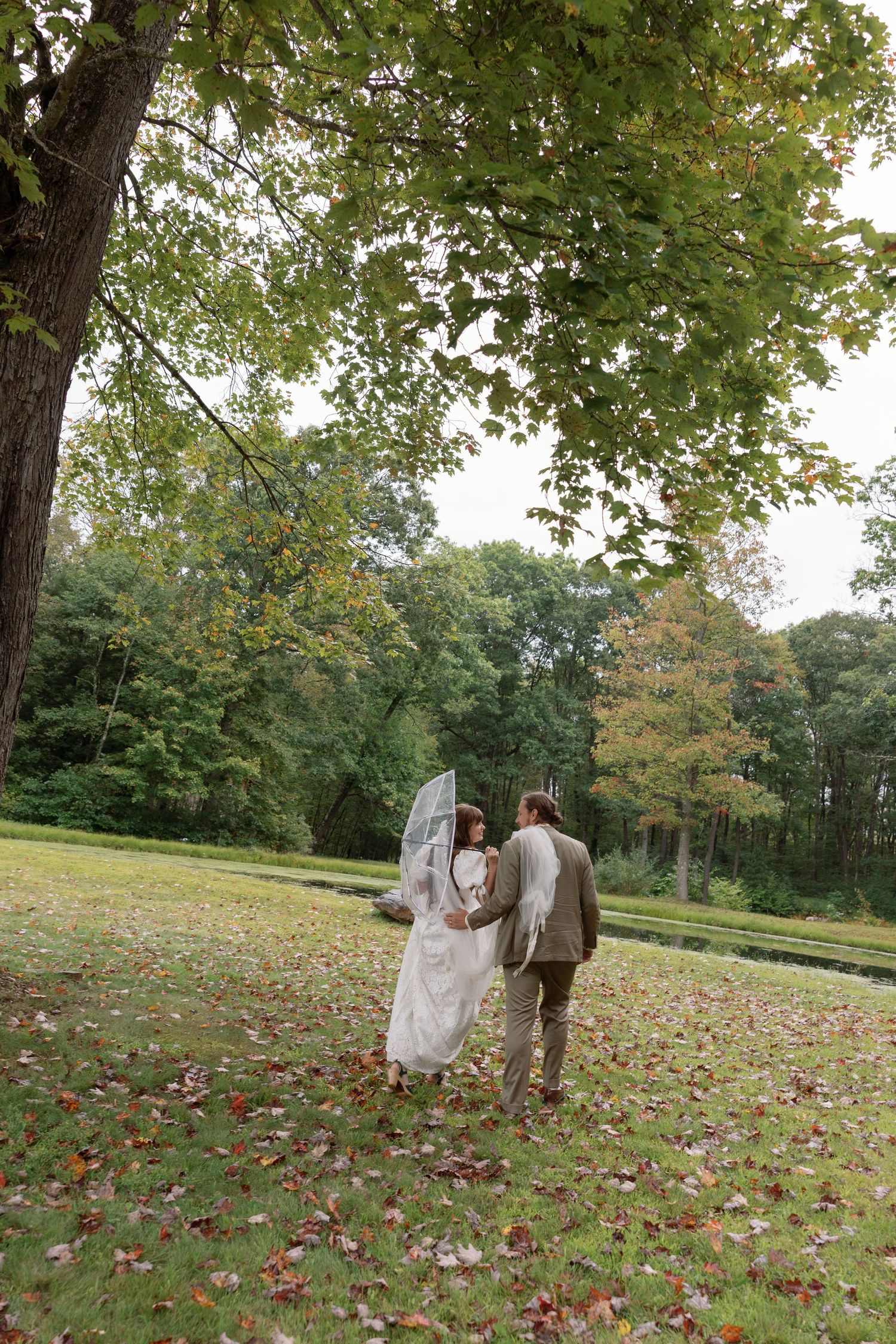 A couple stands together in a scenic autumn park setting with fallen red leaves scattered across the grassy slope.