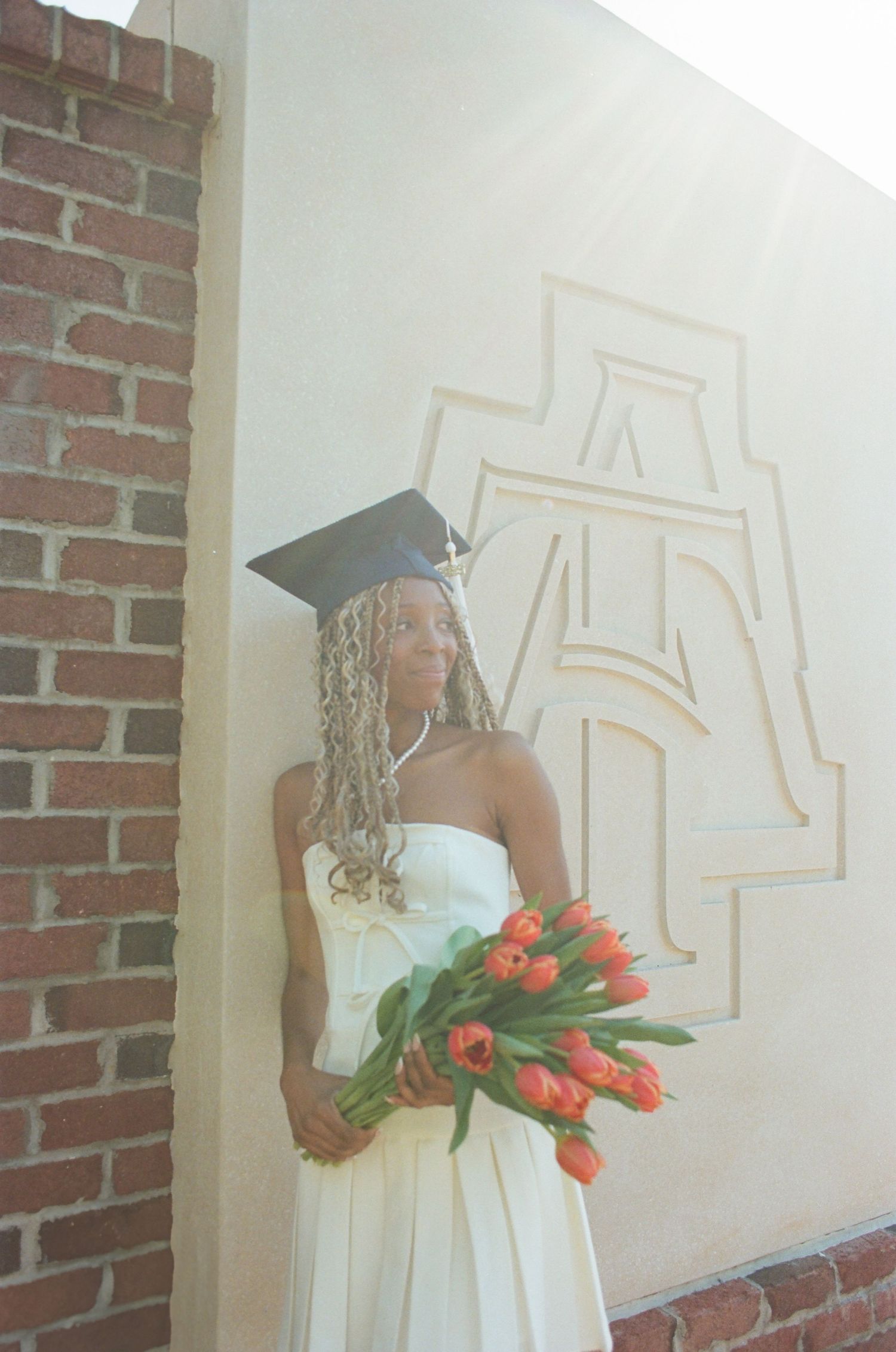 A graduate in white dress holding vibrant red tulips stands by a brick wall with a decorative emblem in the background.