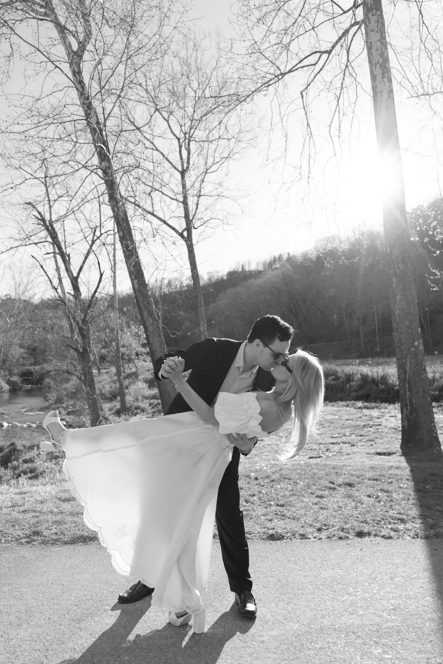 Black and white photo of groom dipping and kissing bride outdoors with sunlight and bare trees in background.