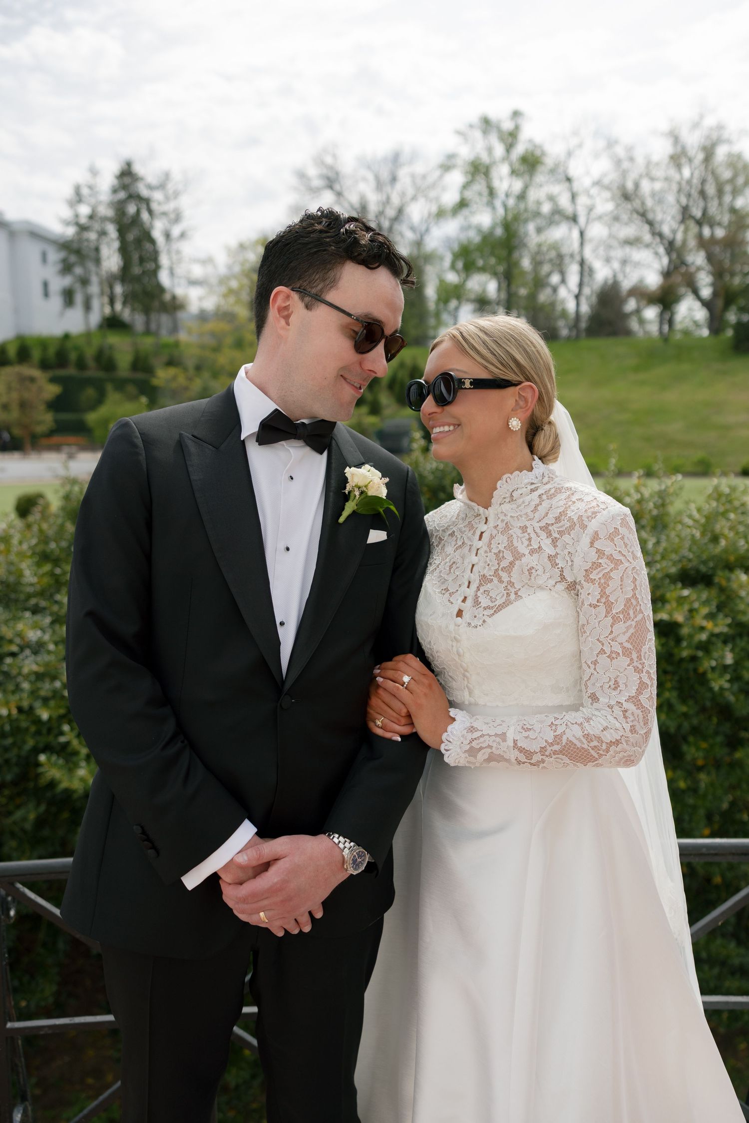A bride and groom in formal wedding attire and sunglasses share a romantic moment outdoors on a sunny day.