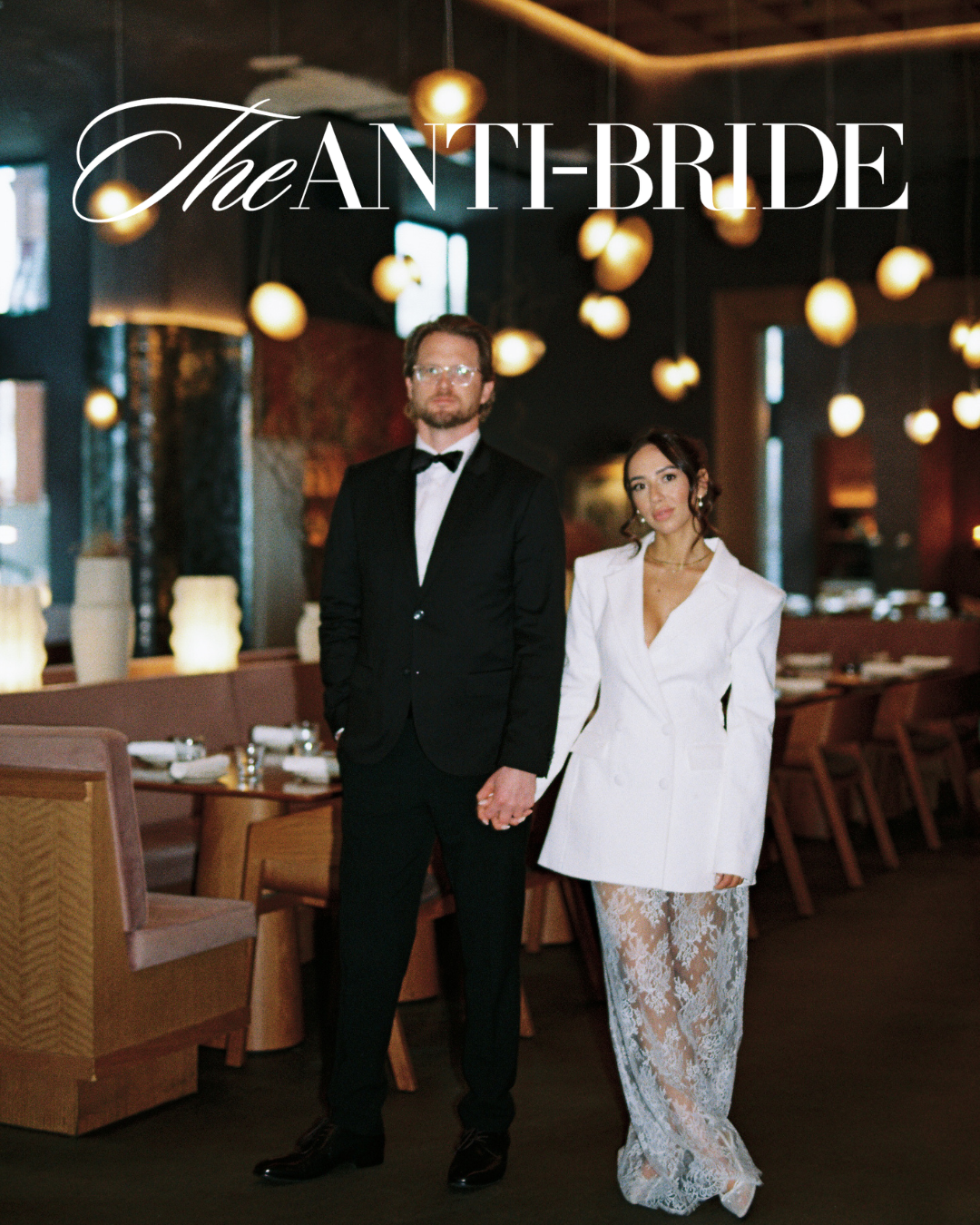 Couple poses in restaurant for The Anti-Bride feature, man in tuxedo and woman in white blazer with lace skirt.