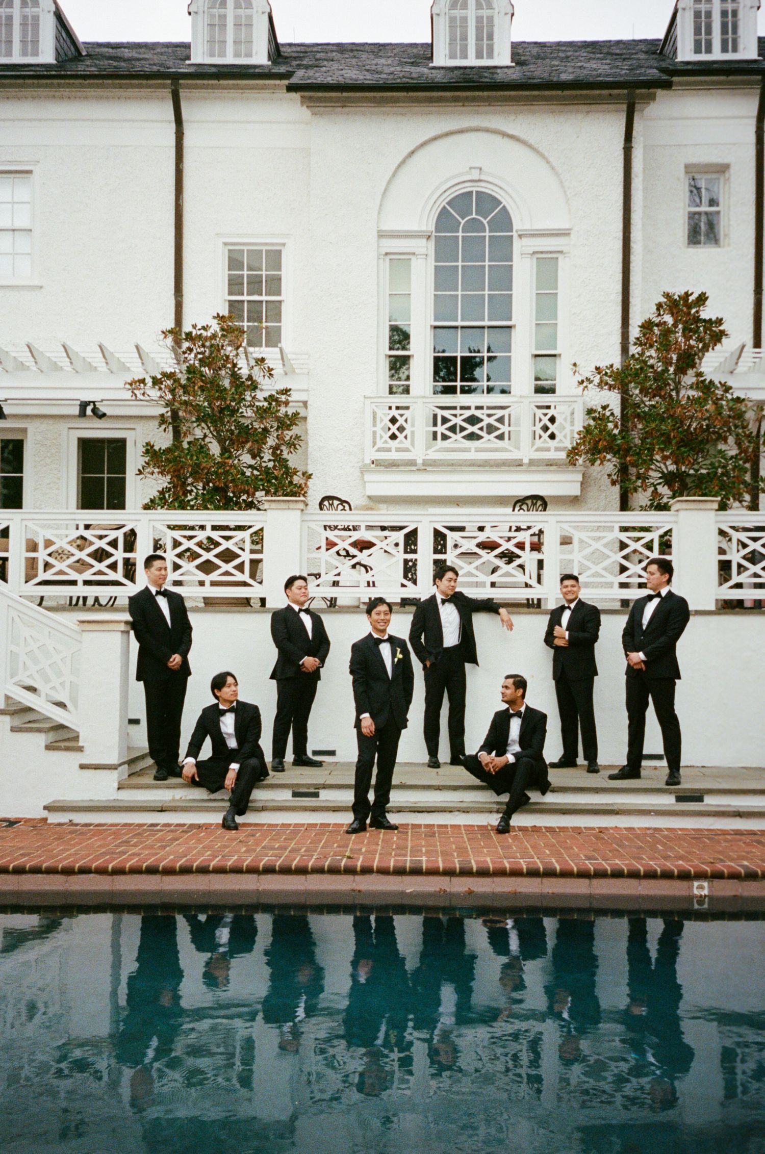 Groomsmen in black tuxedos pose on brick steps beside a pool at an elegant white estate wedding venue.