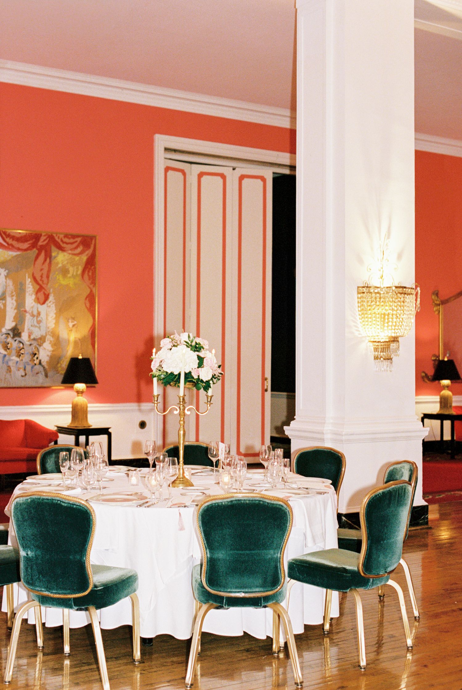 Elegant dining room with coral walls, green velvet chairs, white tablecloth, floral centerpiece, and crystal wall sconce.