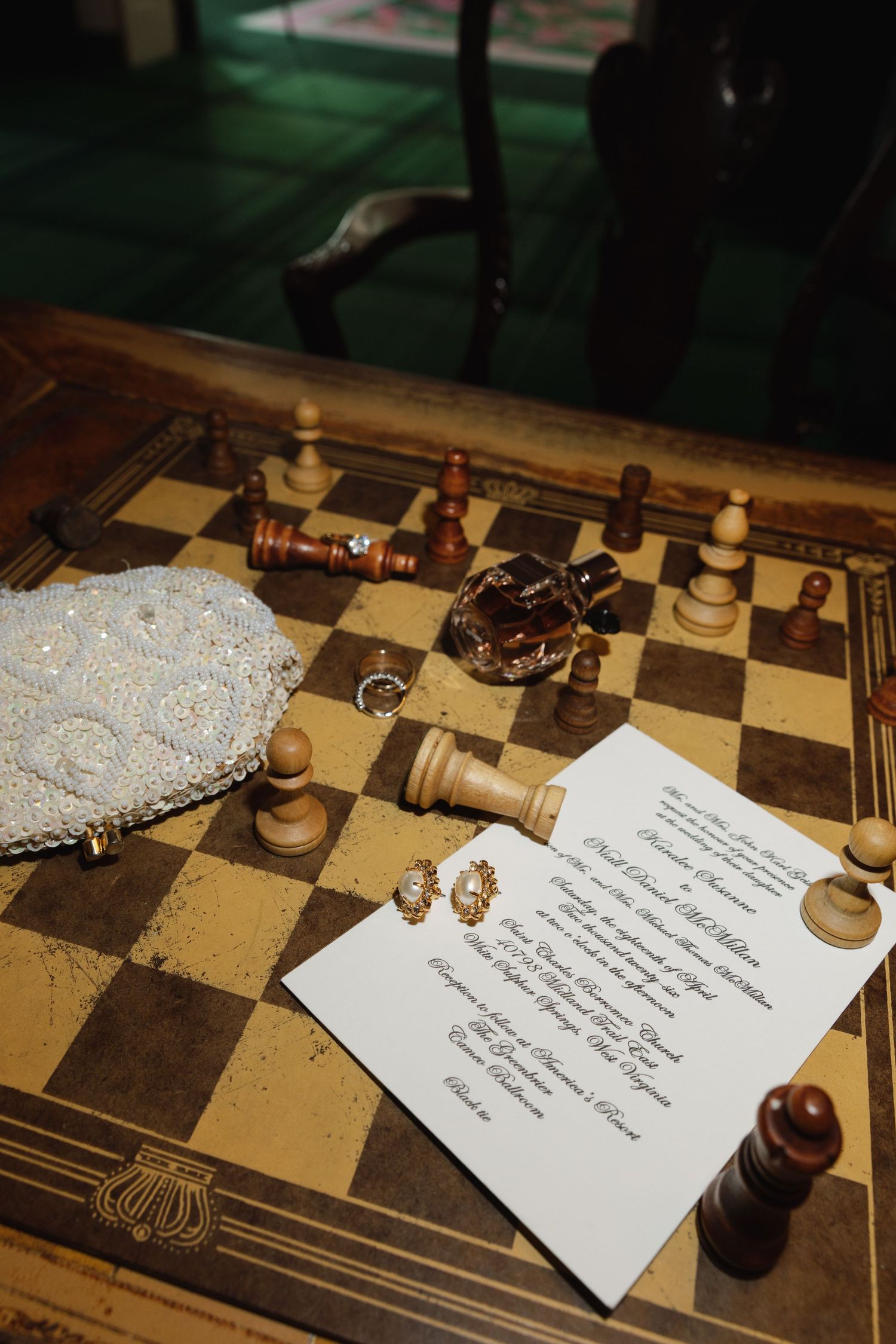 Wedding details on a chess board including an invitation, earrings, perfume, clutch purse, and chess pieces.