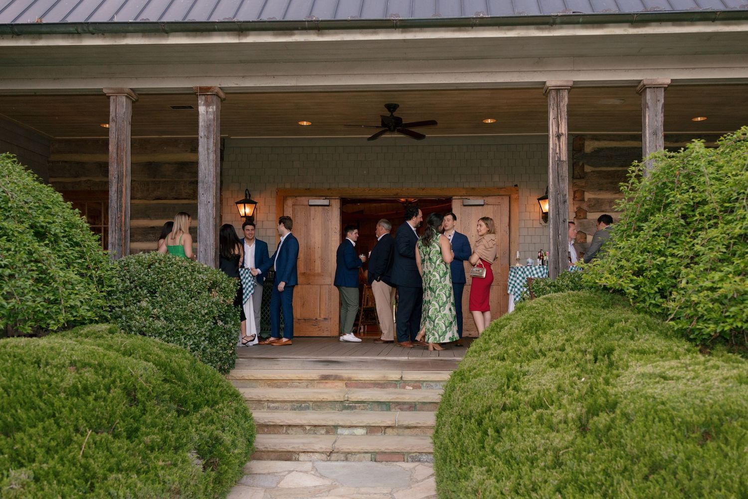 Guests gather on a rustic wooden porch with lush hedges at an elegant outdoor venue event.