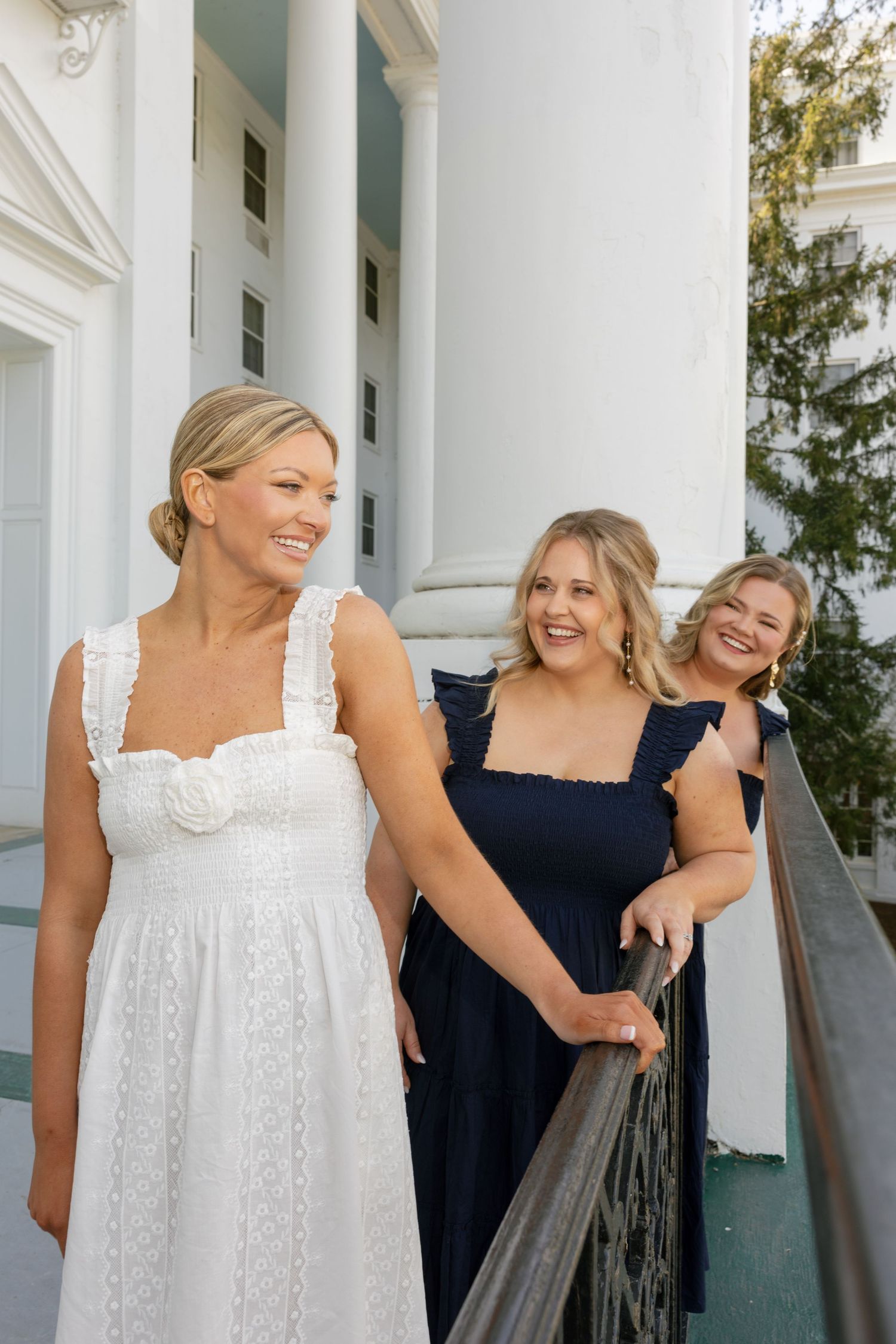 Three smiling women in summer dresses pose on a white columned porch, one in white and two in navy.