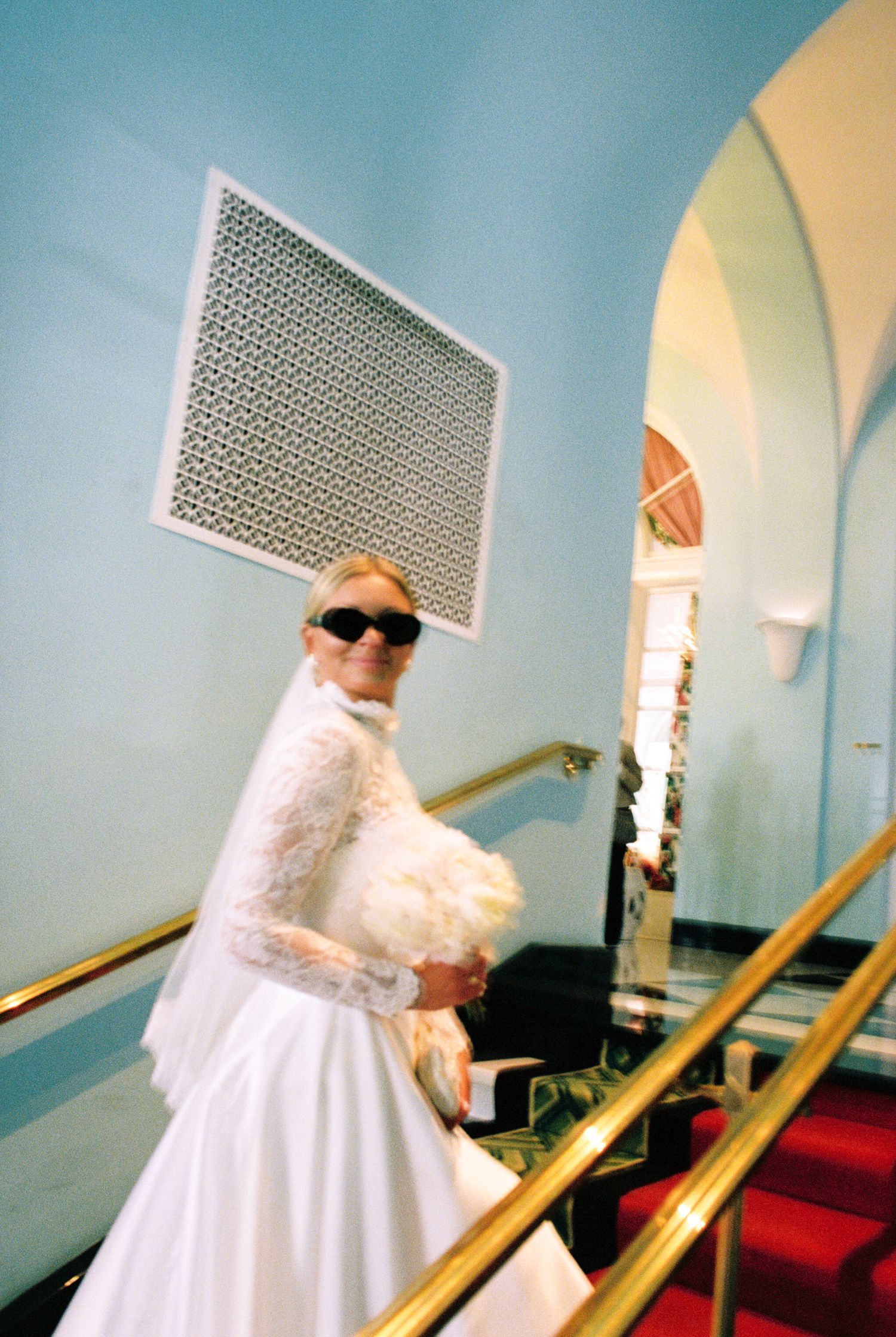 Bride in white lace gown and sunglasses descends grand staircase with gold railing in elegant blue interior.
