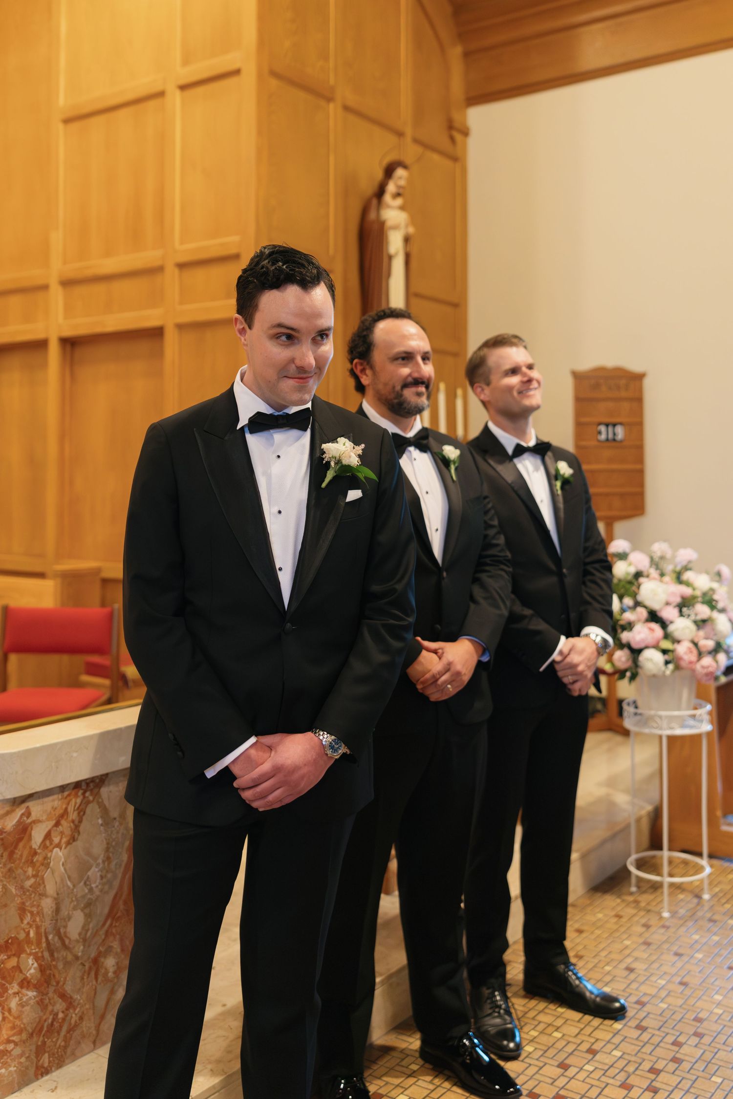 Three groomsmen in black tuxedos with boutonnieres stand at the altar of a church during a wedding ceremony.