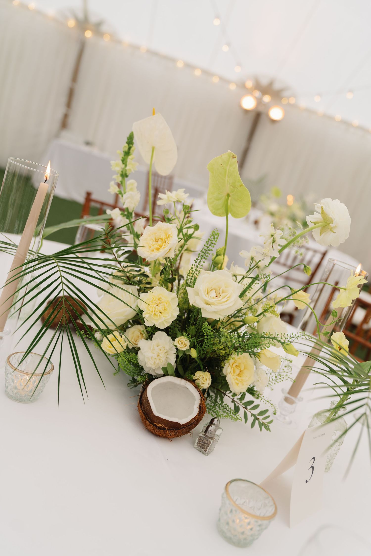 Elegant wedding table centerpiece featuring white roses, palm leaves, and candles in glass holders on a white tablecloth.