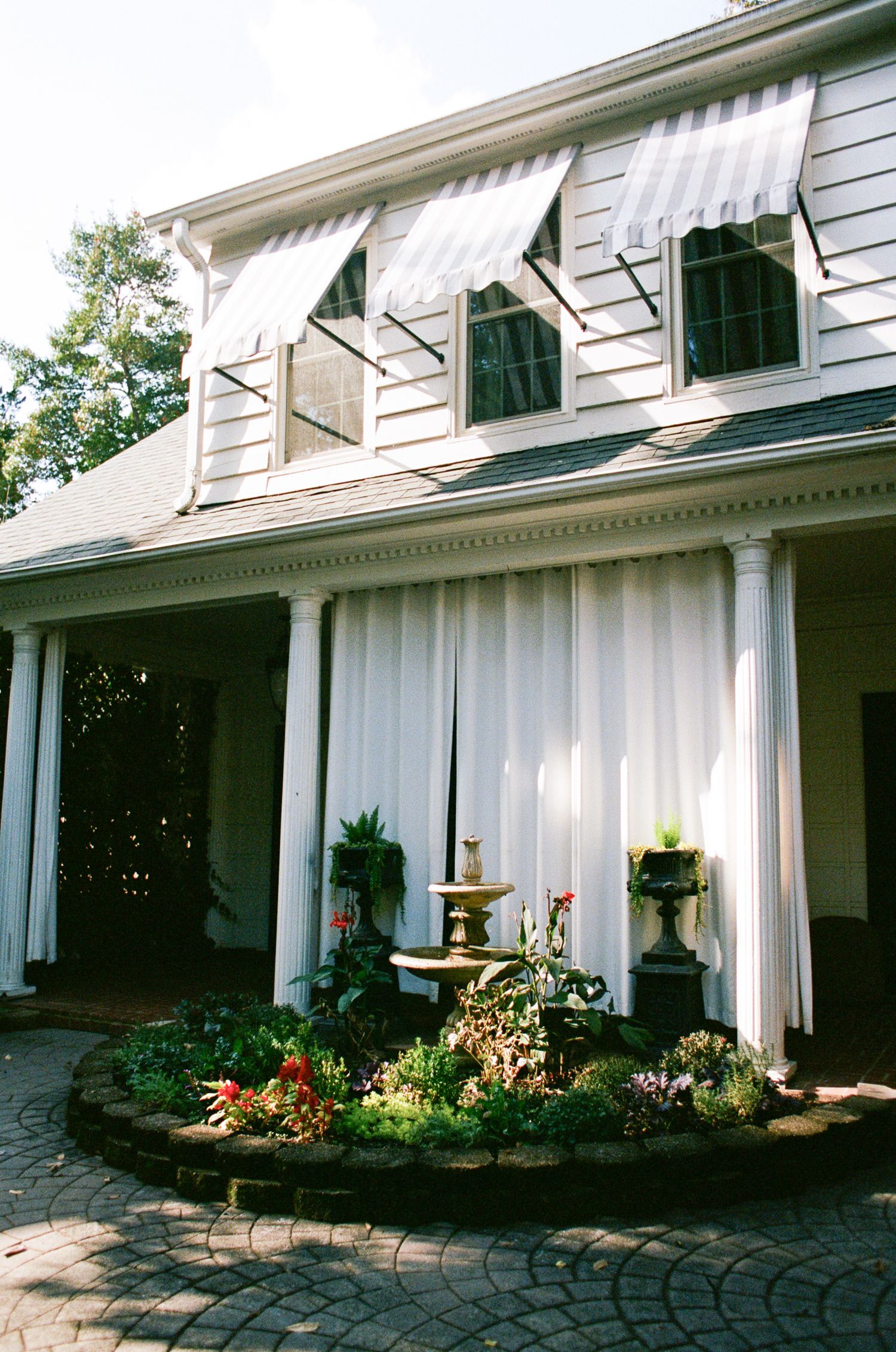 White colonial home with striped awnings, sheer porch curtains, a tiered fountain and colorful flower garden on a brick patio.