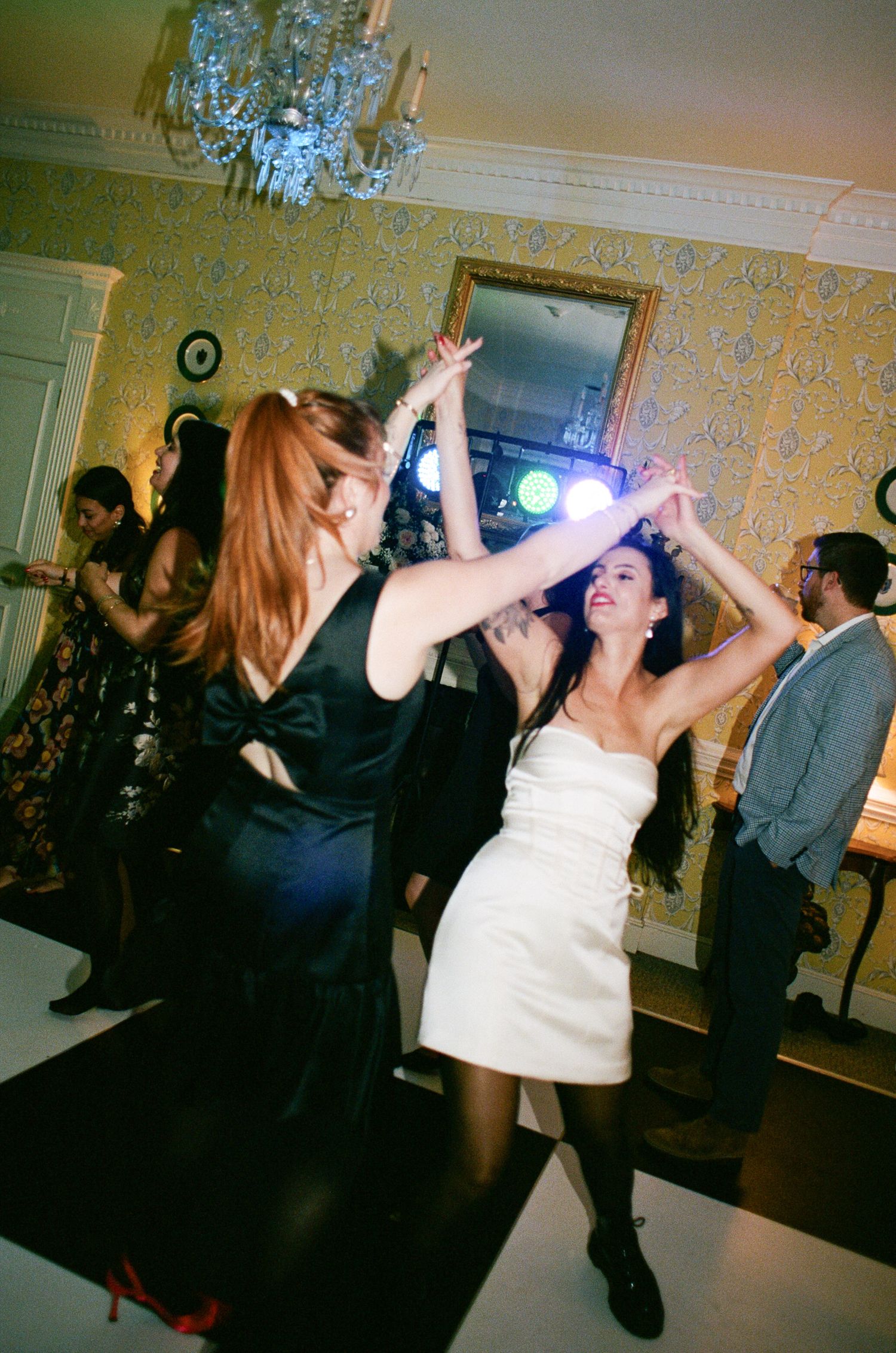 Two women in elegant dresses dance with arms raised at a lively indoor party, with a crystal chandelier and gold mirror in the background.