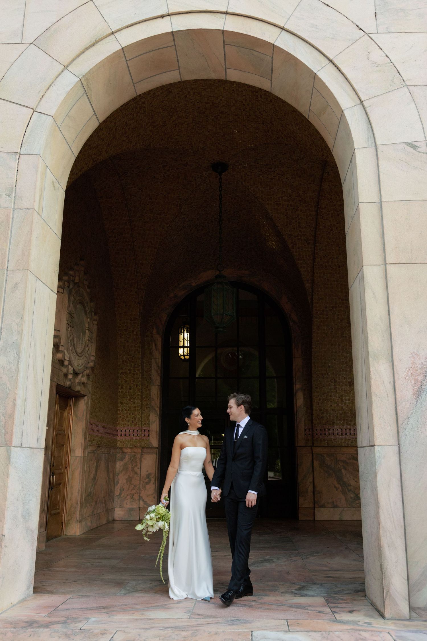 A bride in a white strapless gown holding a bouquet stands beside a groom in a dark suit beneath a grand stone archway.