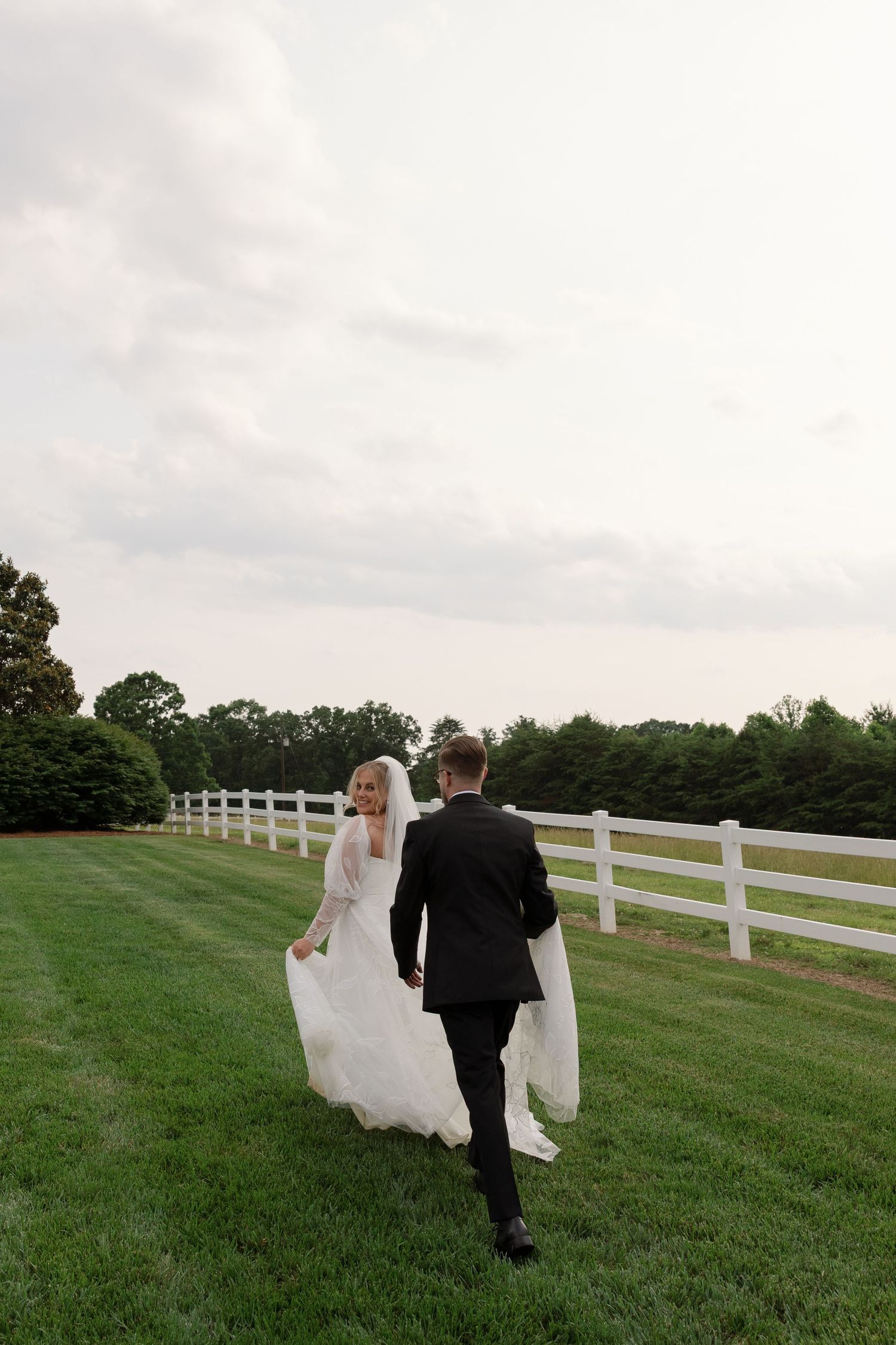 A couple in wedding attire walks along a scenic white-fenced field on their special day.