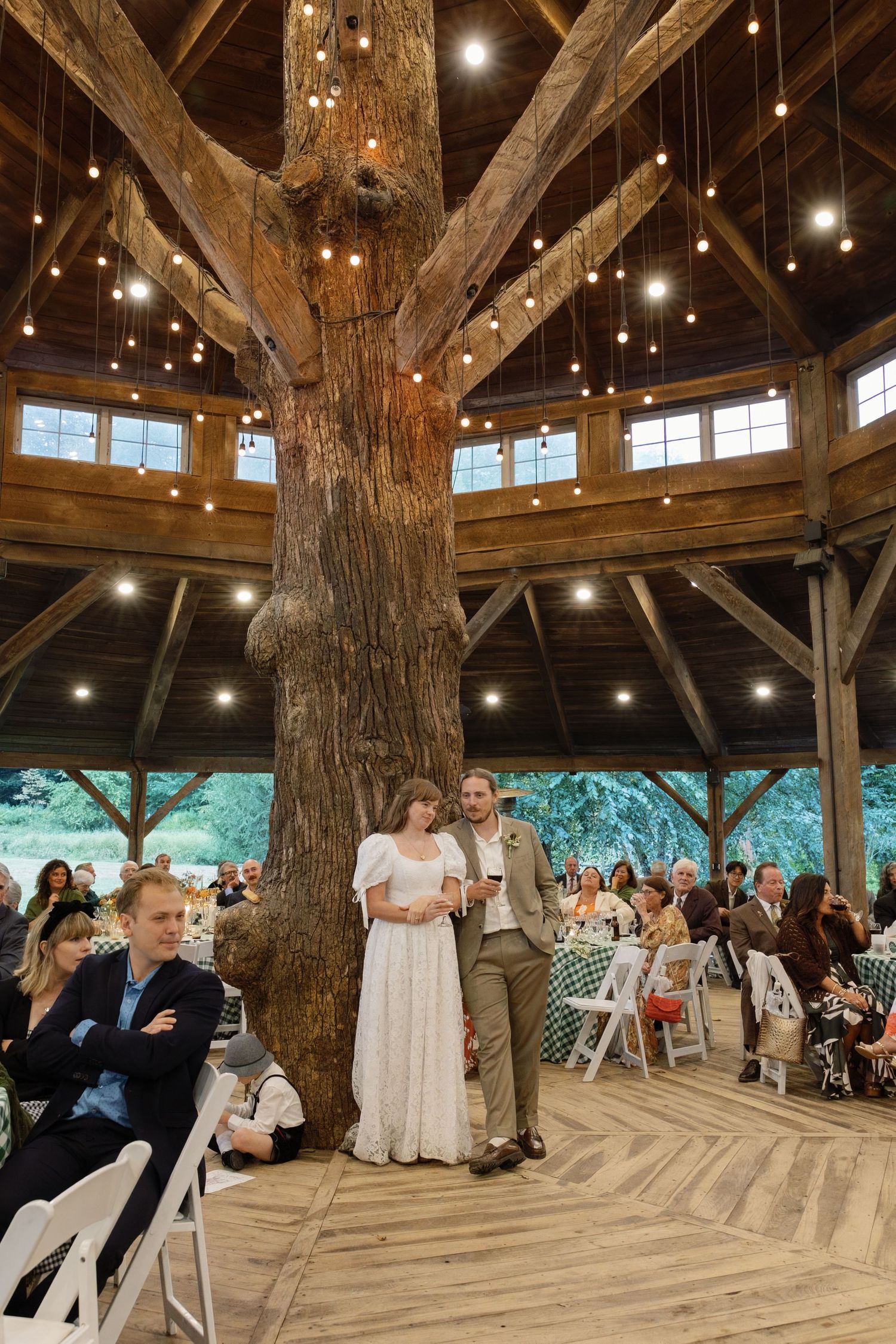 Wedding guests gather in a rustic wooden barn venue decorated with string lights during a ceremony.