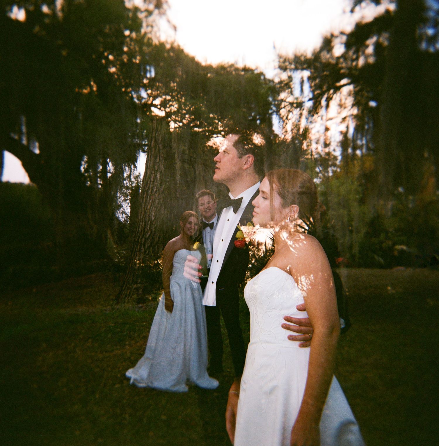 Bride and groom share a romantic moment outdoors at golden hour, with wedding guests visible in the background.