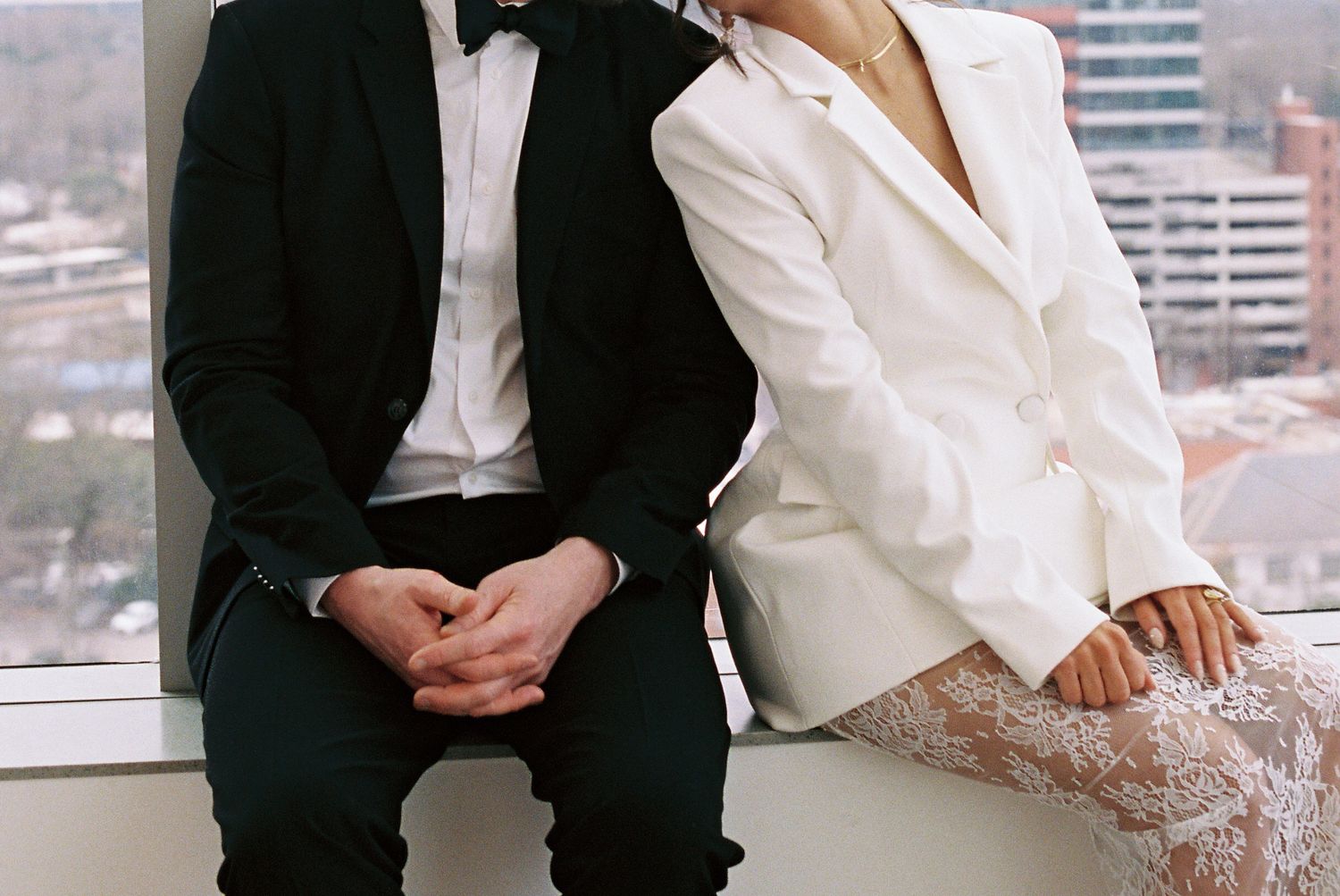 A couple in formal wedding attire, man in black tuxedo and woman in white blazer, sitting together by a window.