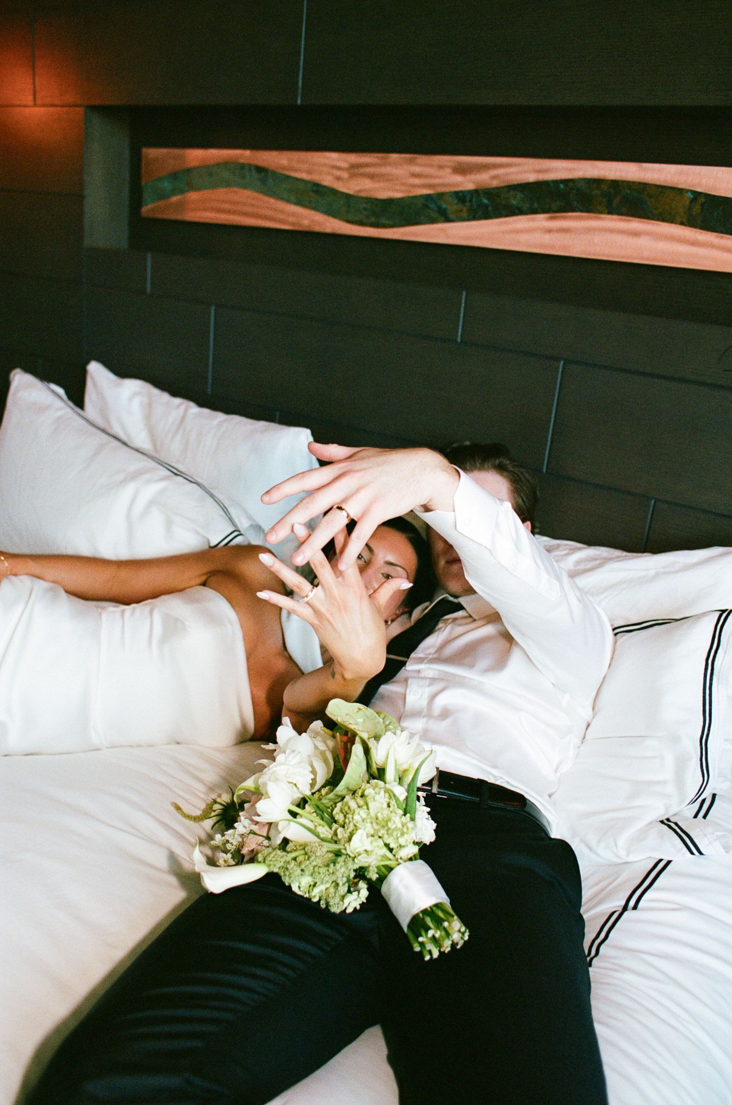 Newlywed couple lying on a white hotel bed, hands raised to show their rings, with a green and white bridal bouquet resting beside them.