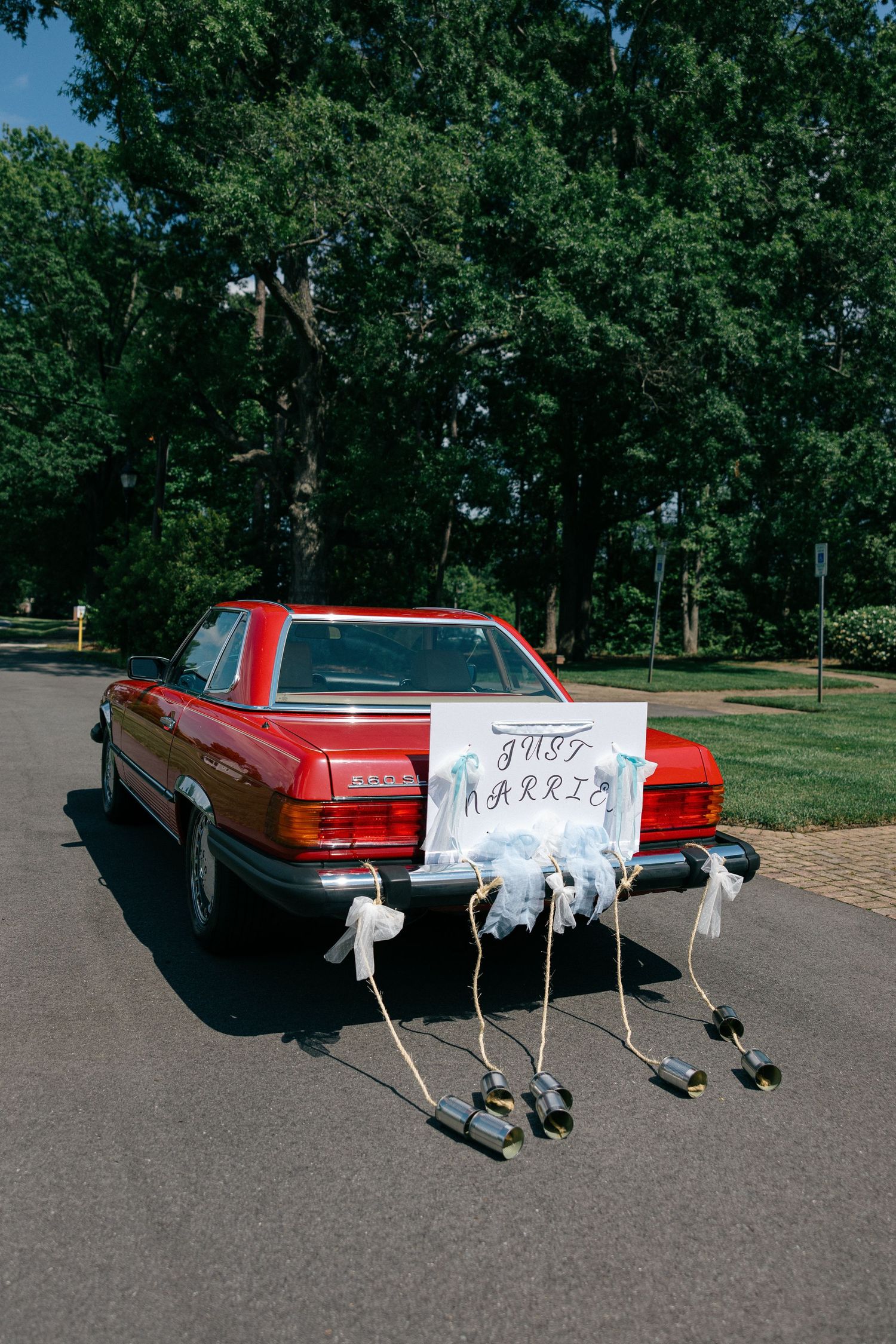 Red vintage convertible with a Just Married sign and tin cans tied to the rear bumper with white ribbons, parked on a tree-lined road.