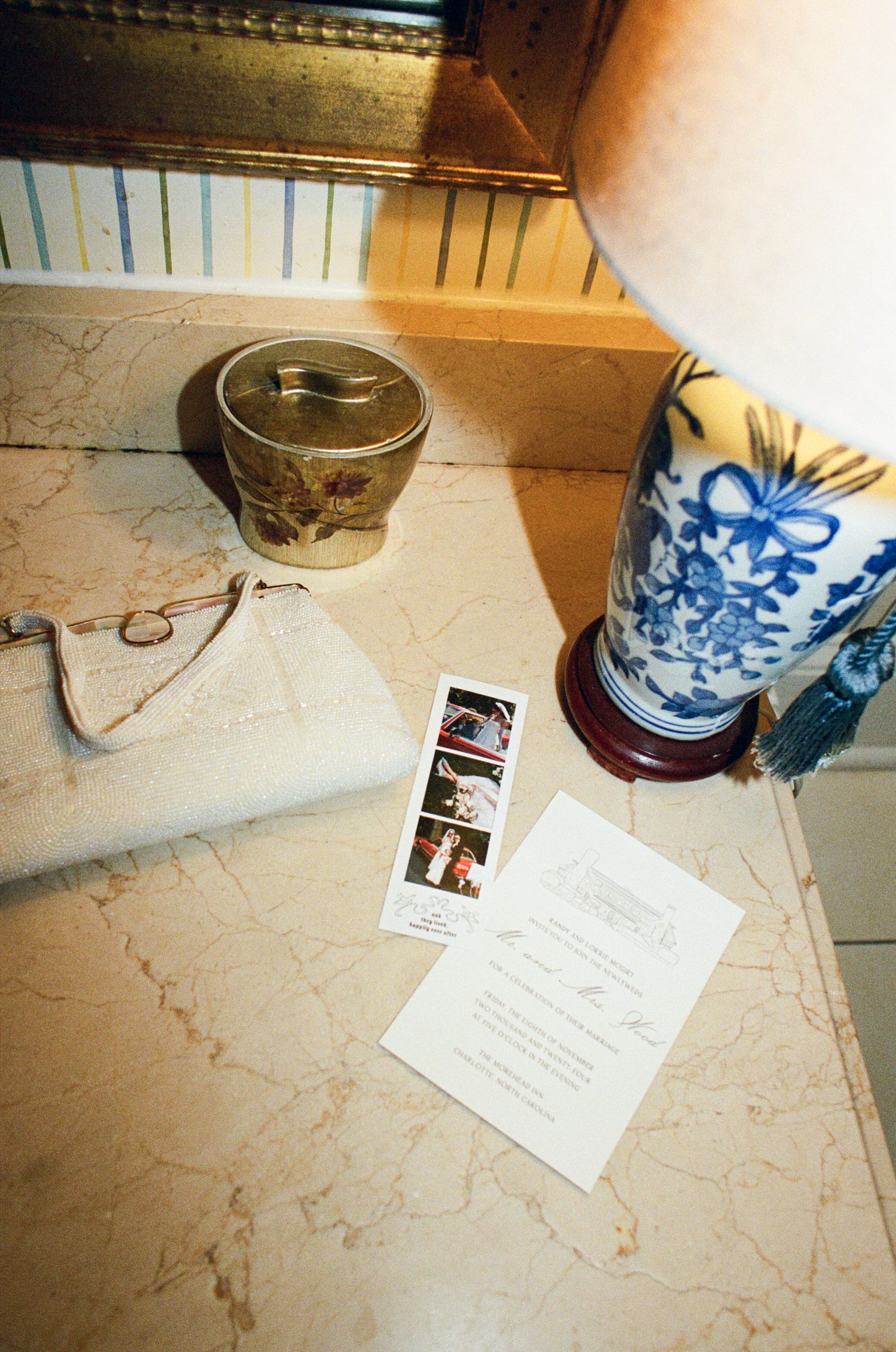 Blue and white porcelain lamp, gold lidded jar, cream clutch purse, photo booth strip and wedding invitation on a marble tabletop.