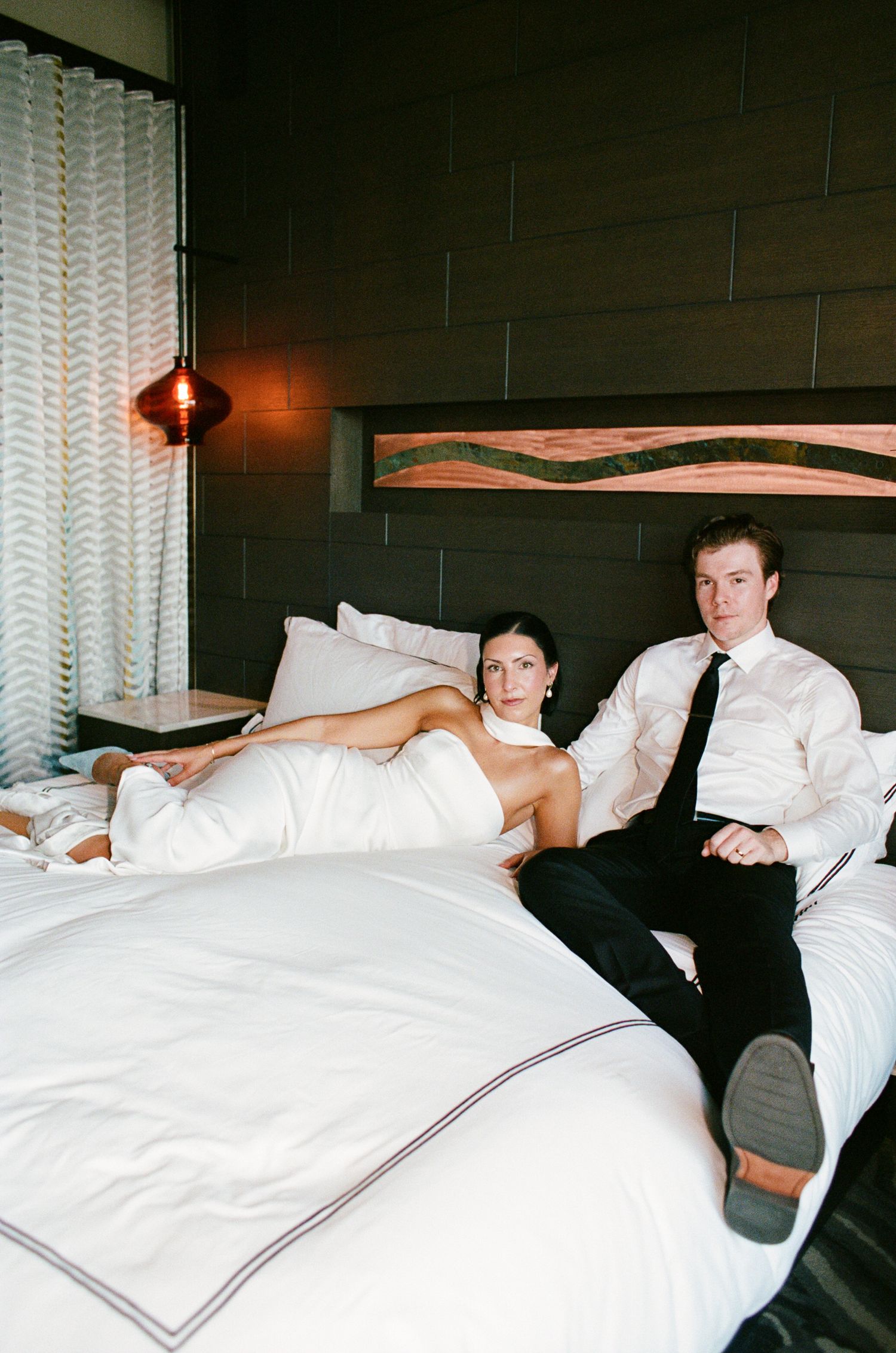 Bride in a white gown reclines on a white hotel bed beside a groom in a white shirt and black tie, in a modern room with wood paneling.