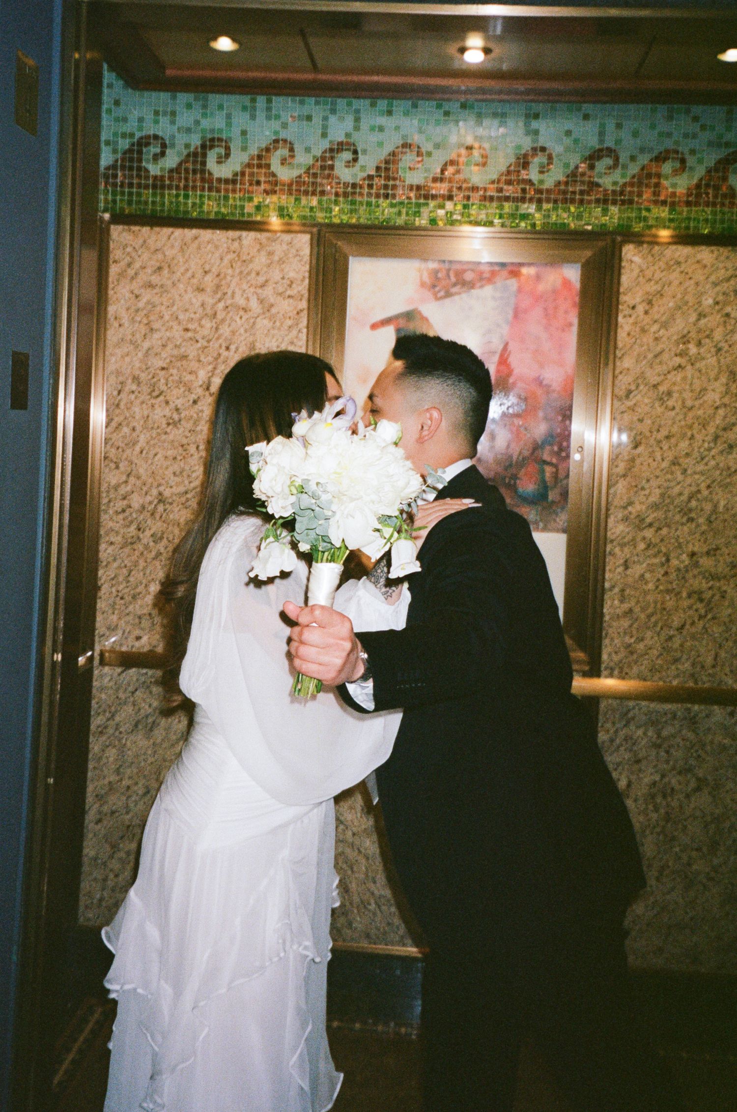 A bride in a white wedding dress and veil holds a bouquet while walking through a dimly lit hallway.
