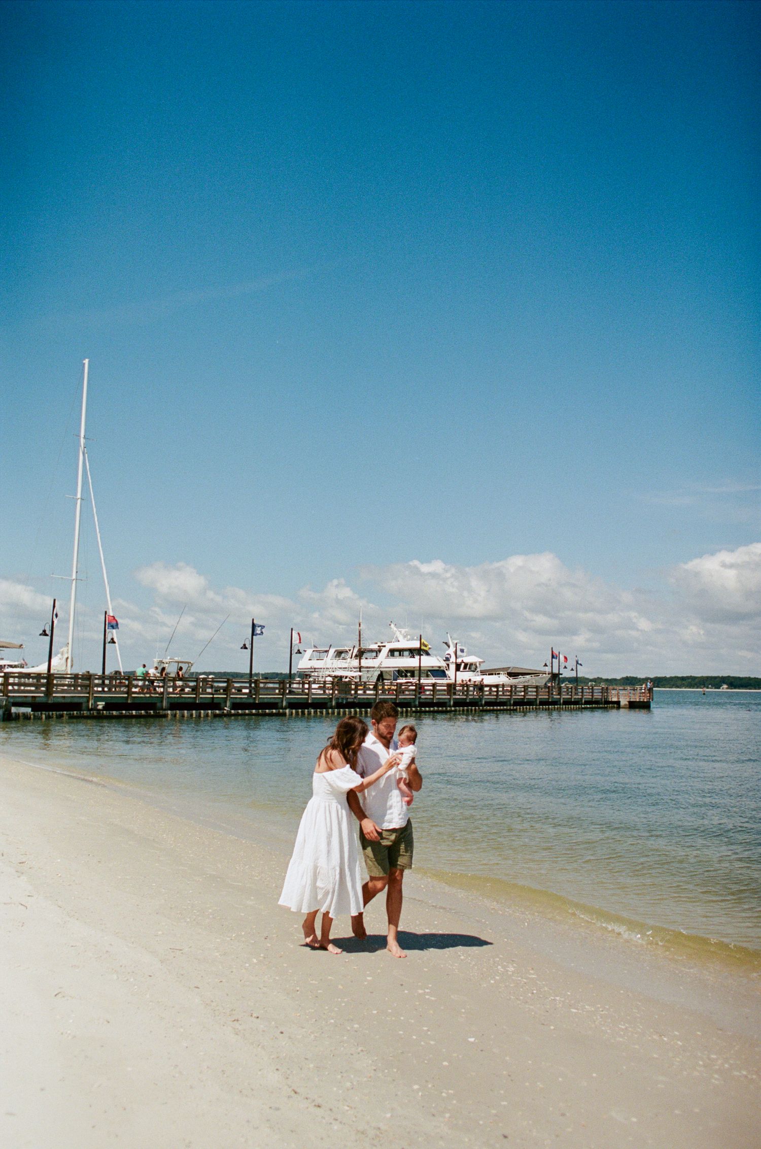 A panoramic view of people walking on a sandy beach with boats and docks in the distance under a bright blue sky.