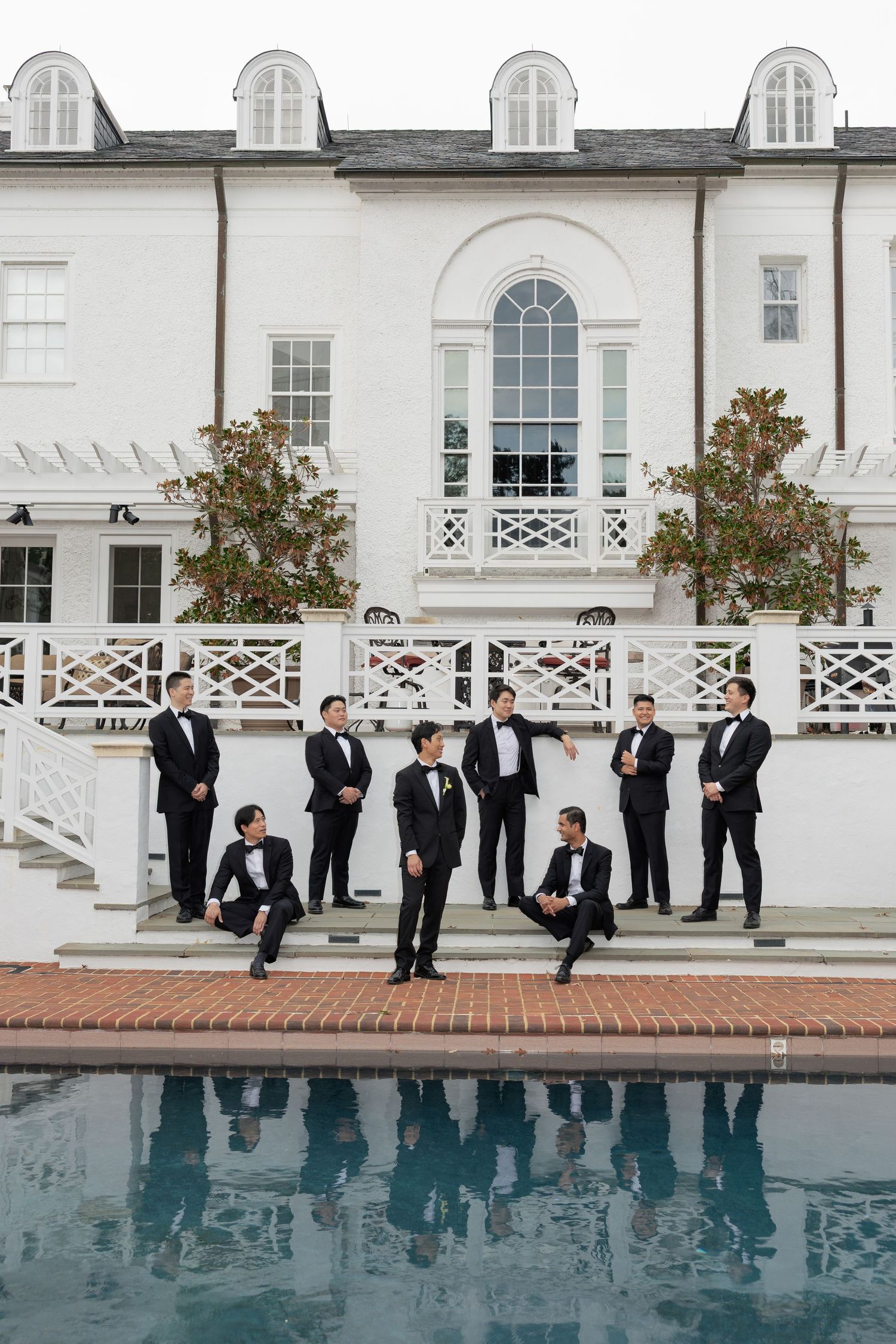 Groomsmen in black tuxedos pose along a white railing by a swimming pool at an elegant venue.