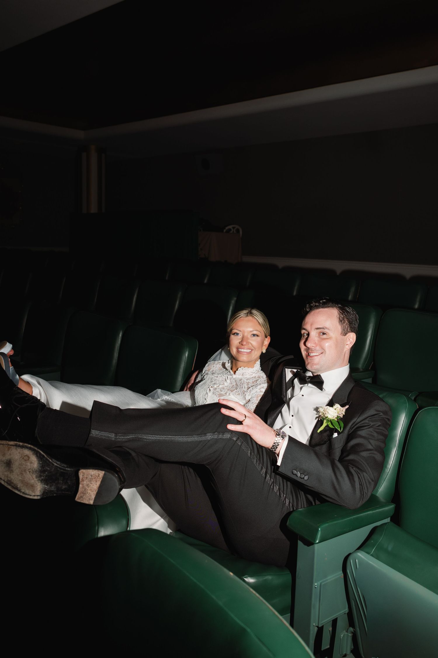 Bride and groom in formal attire laughing together in green theater seats, groom with legs up, bride holding a drink.