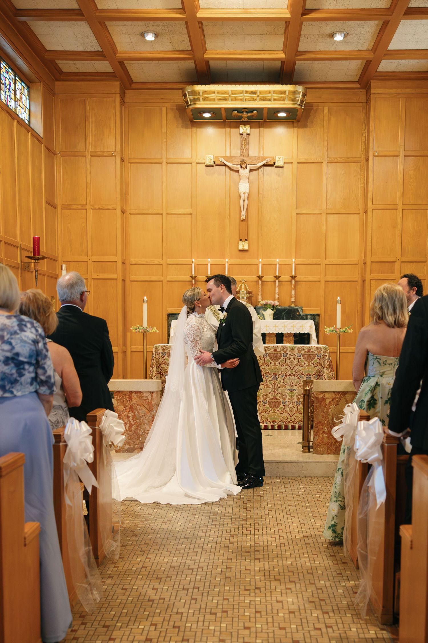 Bride and groom share first kiss at altar in elegant wood-paneled Catholic chapel with crucifix and candles.