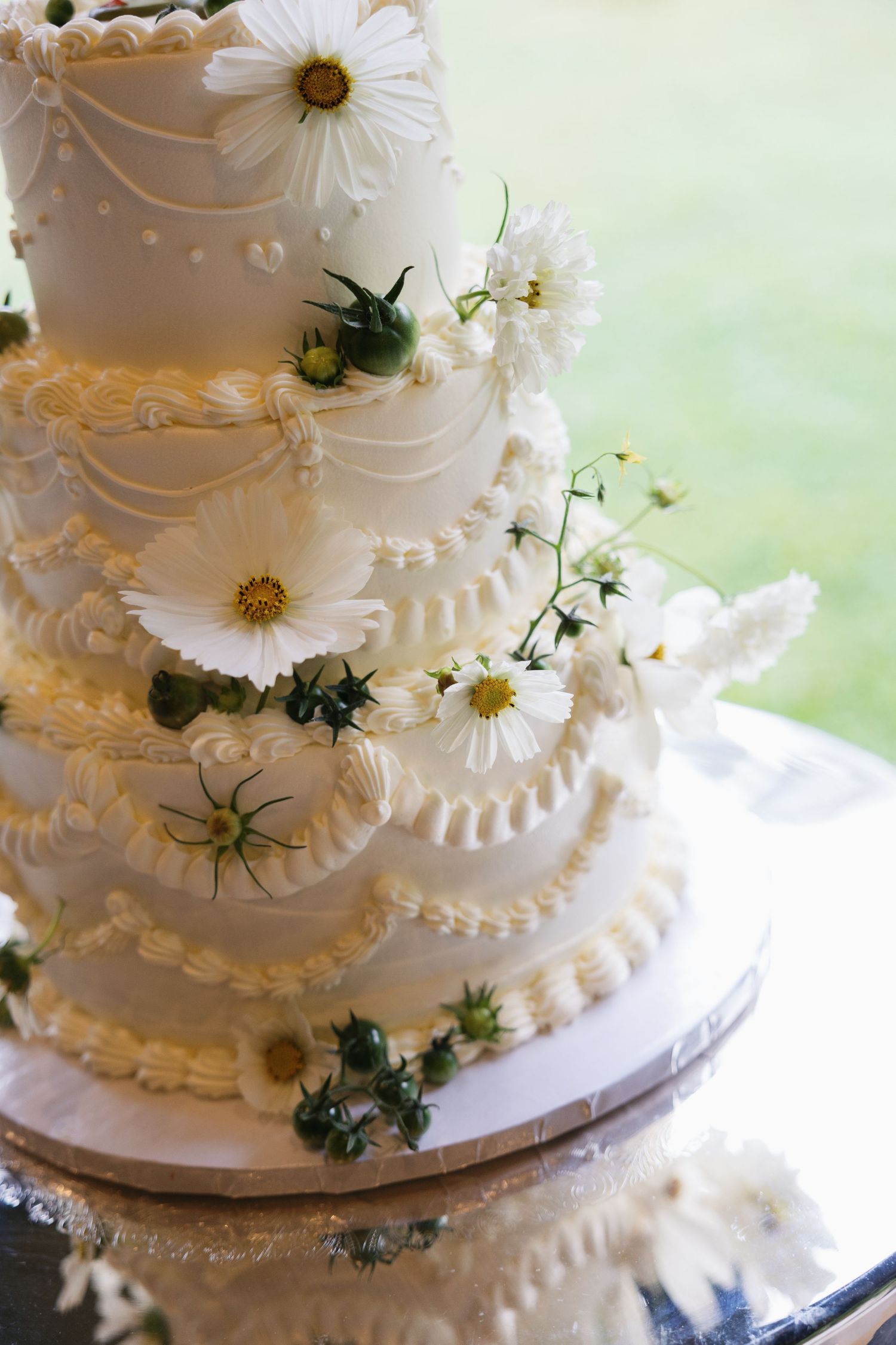 Close-up details of an elegant white wedding cake decorated with delicate piping, fresh daisies and greenery accents.